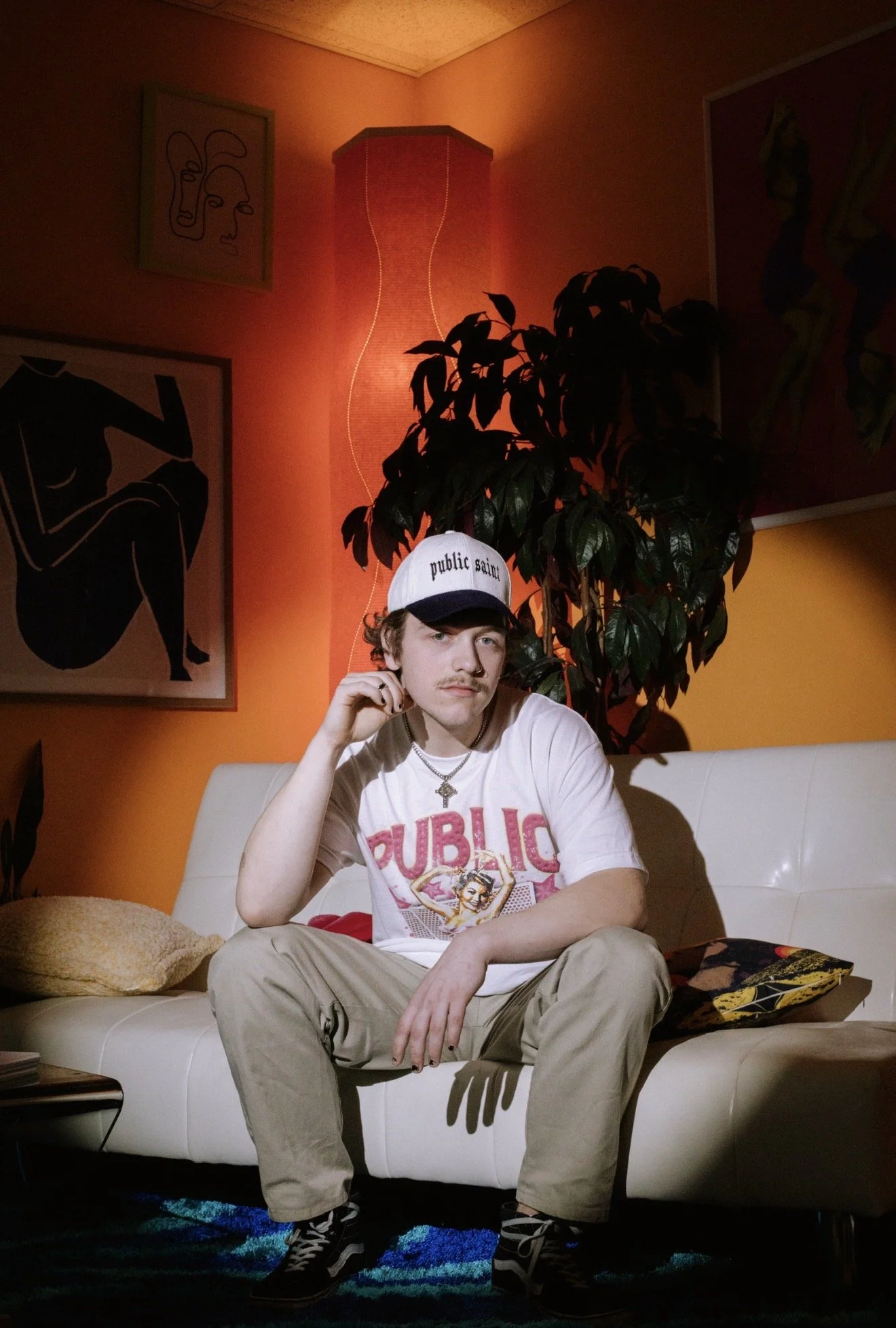 A young man with a mustache and curly hair sits on a white couch in a room with orange walls and colorful abstract art. He wears a white t-shirt with a graphic print, tan pants, a baseball cap that says "public saint," and a necklace with a cross. Th