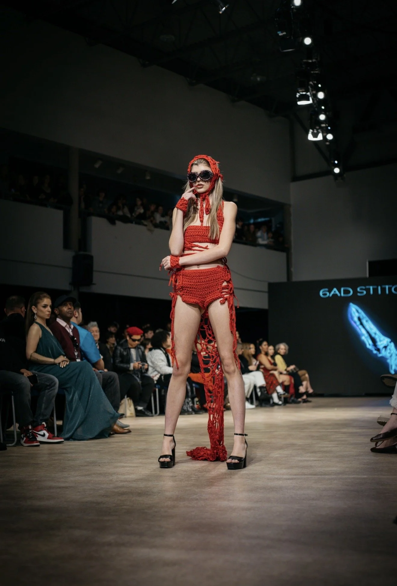 Model walking on runway in red crochet top and shorts with matching accessories, wearing sunglasses, in a fashion show.