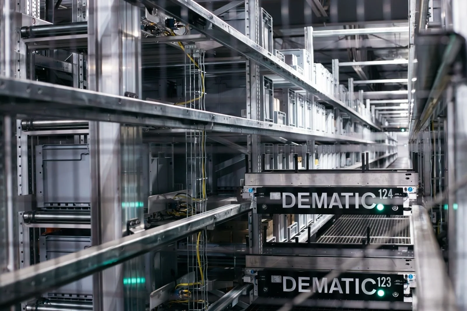 Inside a server room with multiple metal shelves holding electronic equipment and network cables, with a robotic system labeled 'DEMATIC 124' and 'DEMATIC 123' in the foreground.