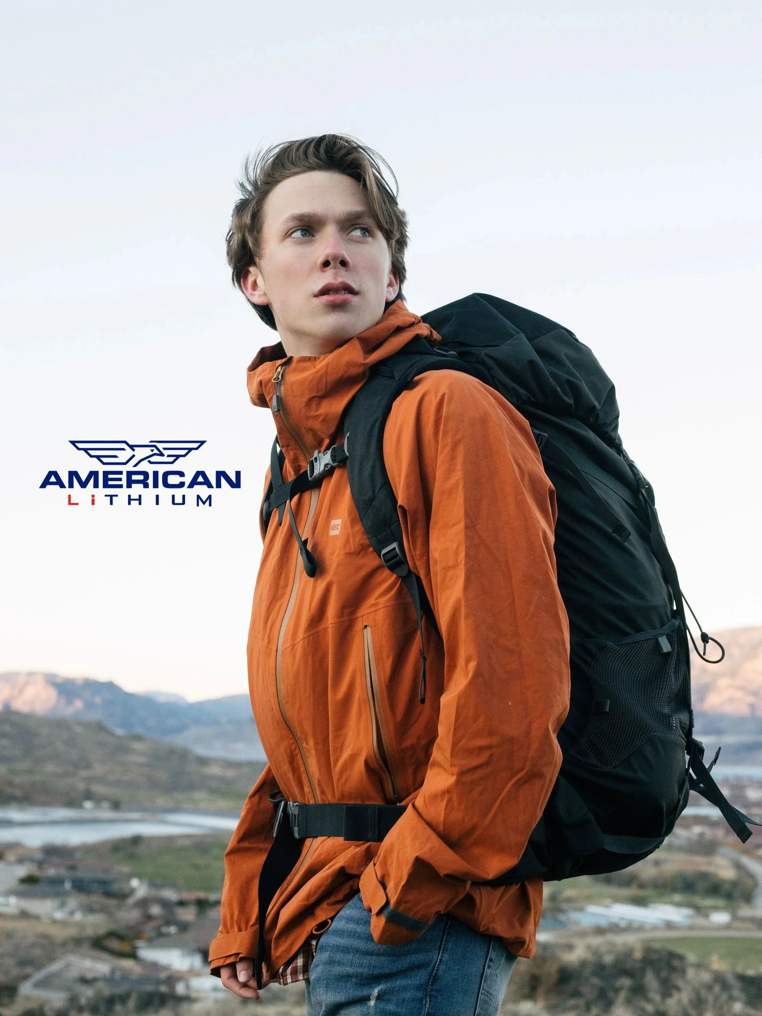 Young man with brown hair wearing an orange jacket and backpack, standing outdoors with hills and water in background. Wearing MEC outdoor clothing on a hiking trail in the mountains that was shot for American Lithium, a mining company in Nevada.