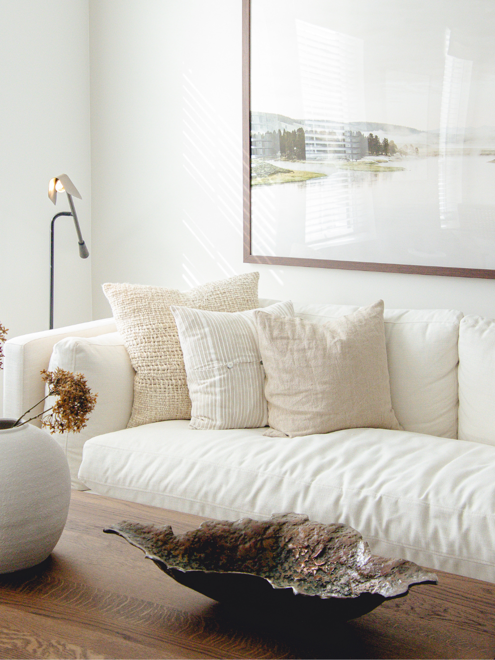 Living room with a white sofa, beige cushions, a wooden coffee table with a decorative bowl, a tall floor lamp, and a large framed landscape photograph on the wall, illuminated by natural light.