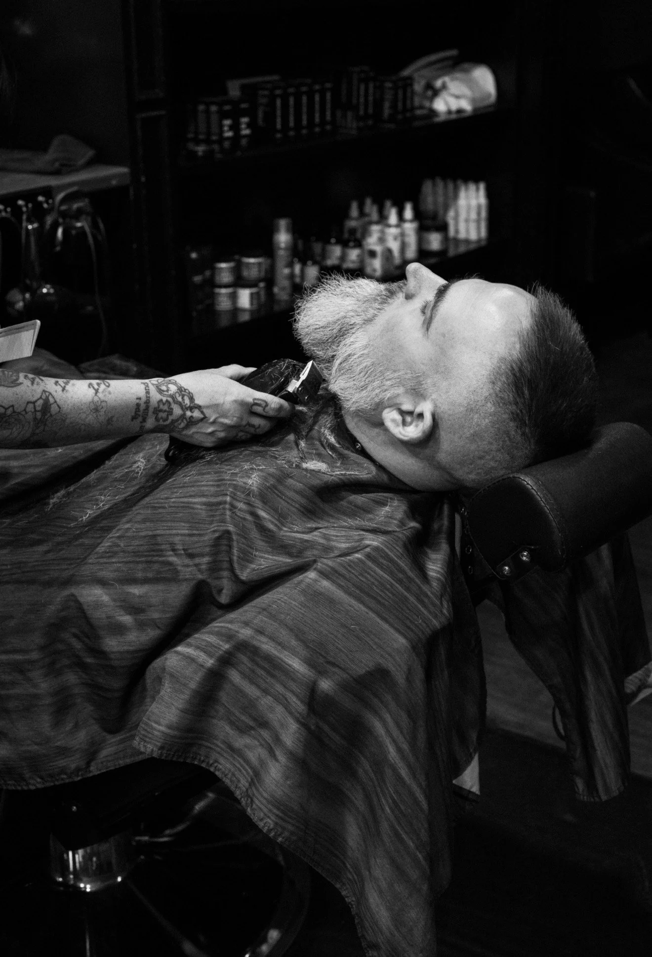 A man with a beard is lying back in a barber's chair with his eyes closed, receiving a haircut or shave, with a barber using clippers on his beard. The scene is in a barbershop with shelves of grooming products in the background.