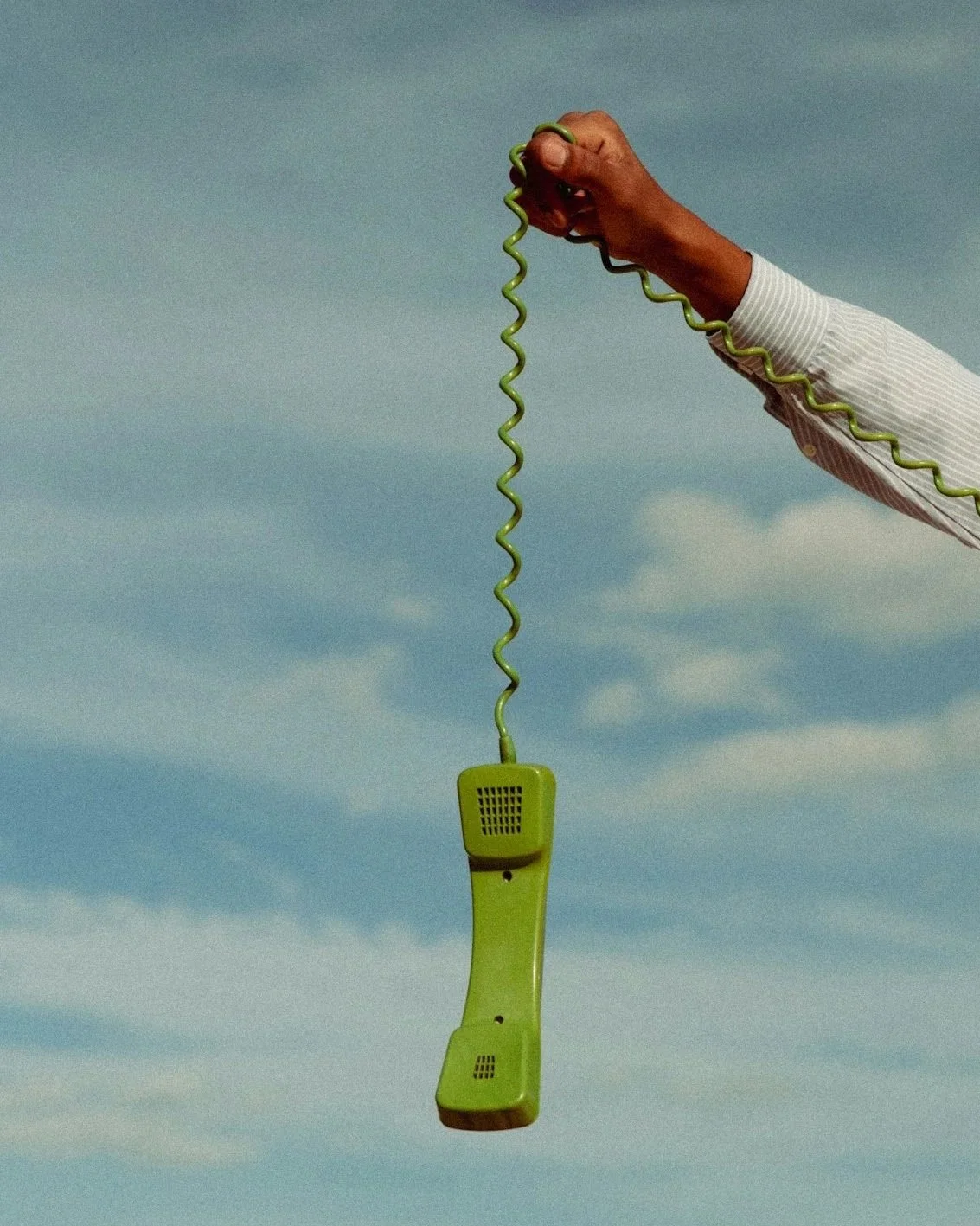 A person in a striped shirt holds a vintage green landline telephone by its cord against a cloudy sky.