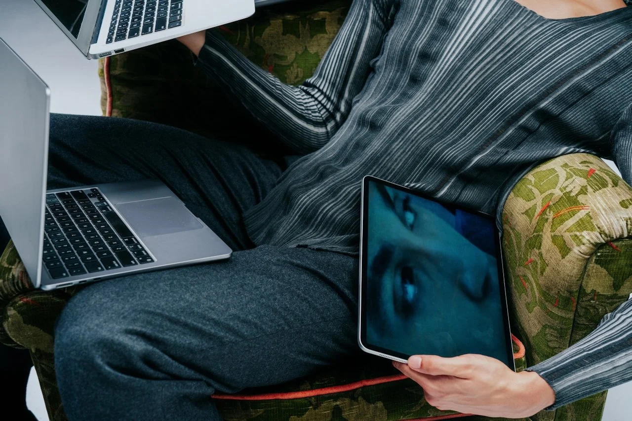 Person sitting on a vintage floral patterned couch using multiple electronic devices including a laptop, a tablet with a woman's face on the screen, and a smartphone, all within a living room setting.