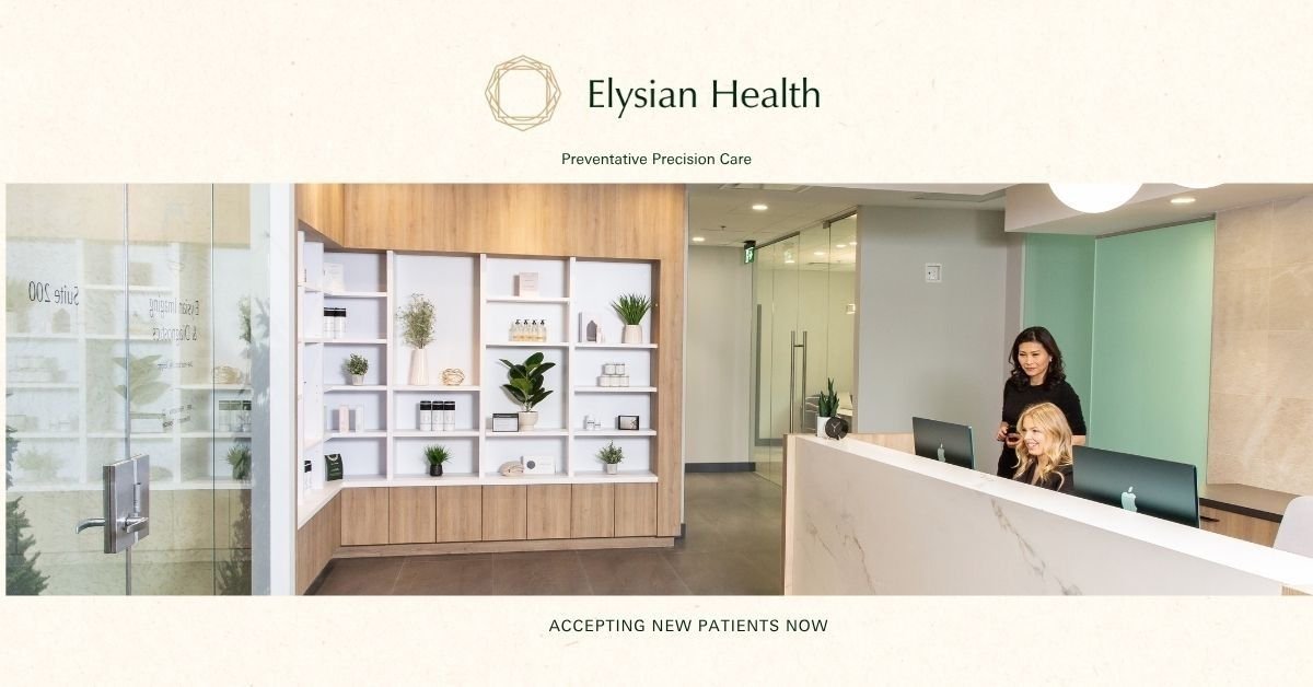 Reception area of Elysian Health, a medical office with a wooden and white desk, two staff members working, and decorative shelving with plants and decor