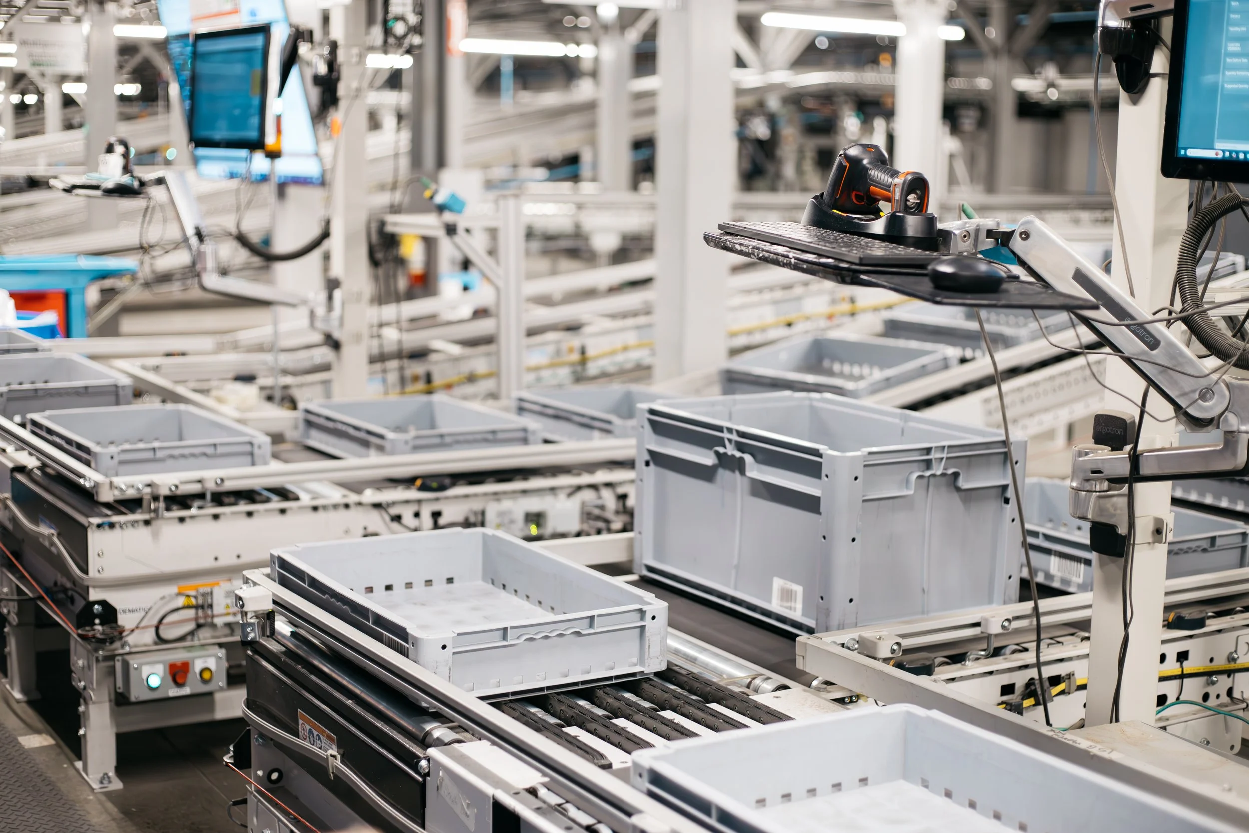 Interior of an automated warehouse or manufacturing facility with conveyor system, empty plastic bins, and computer monitors.