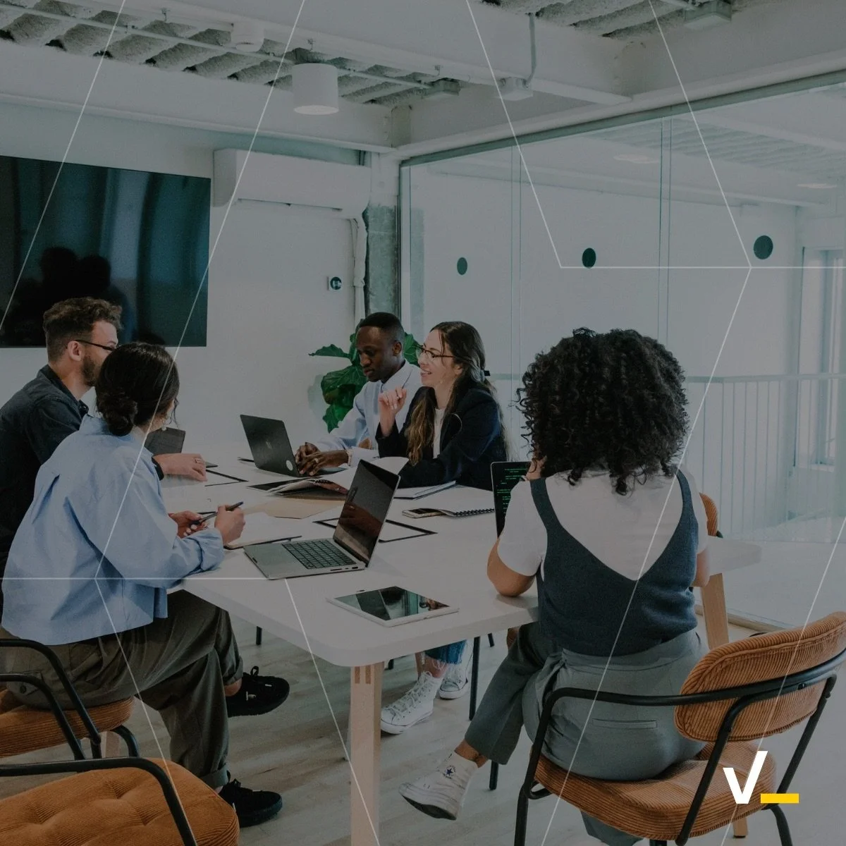 A diverse group of six people sitting around a conference table in a modern office, engaging in a meeting with laptops and notes. The room has a large window, a TV on the wall, and is well-lit with natural light.