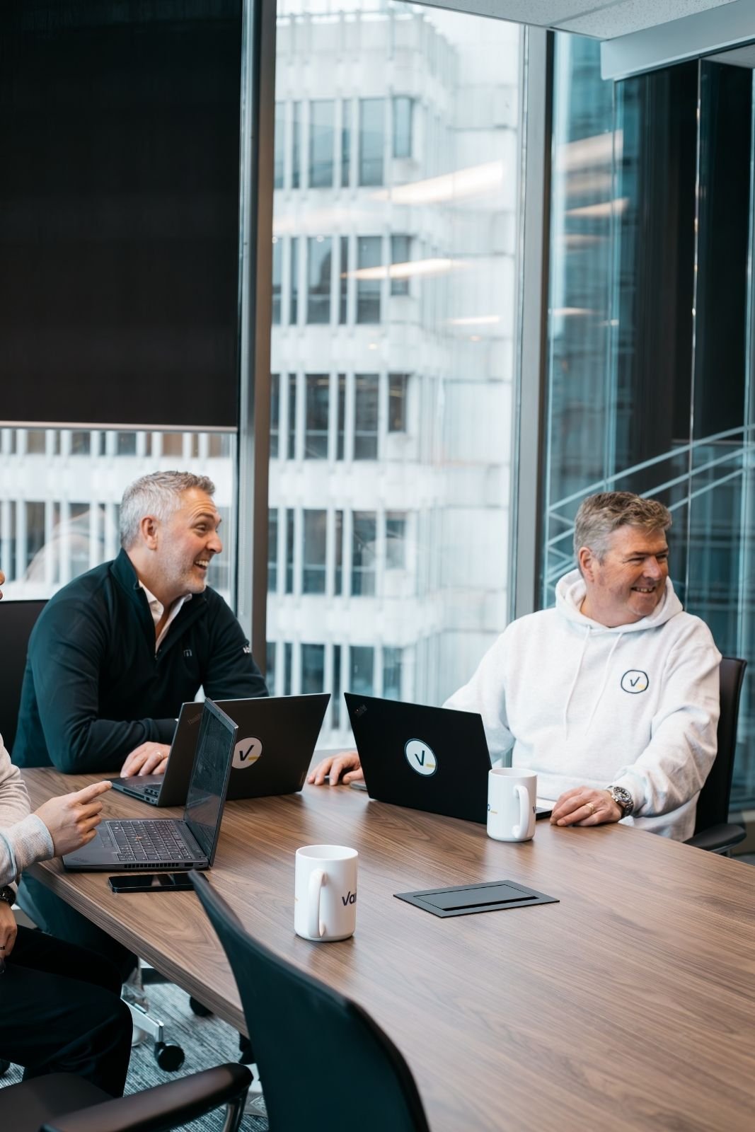 Three men sitting at a conference table with laptops, smiling and talking, in a modern office with large windows showing a cityscape.