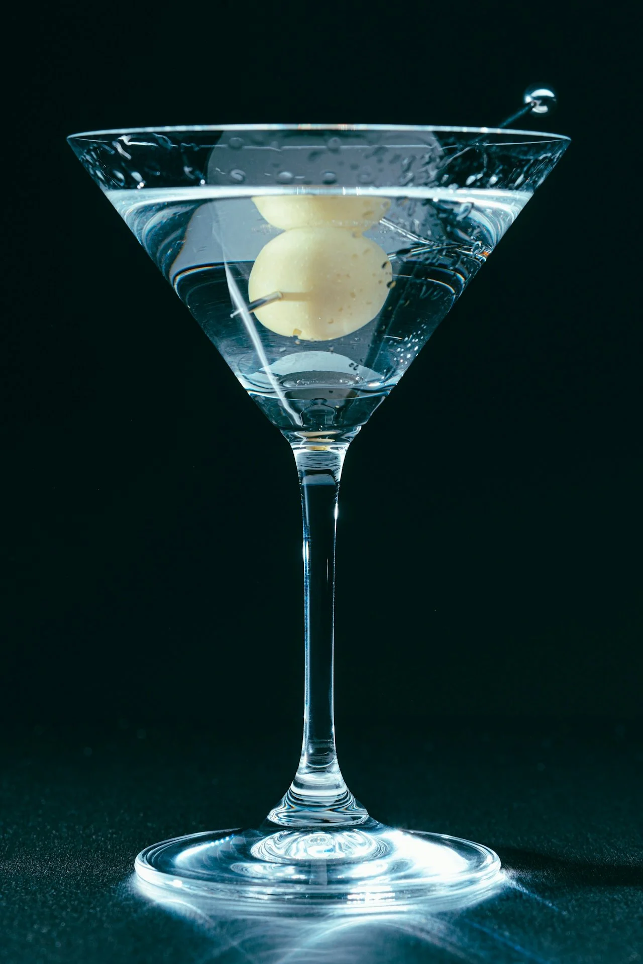 A classic martini glass filled with a clear cocktail, garnished with an olive on a cocktail pick, against a black background.