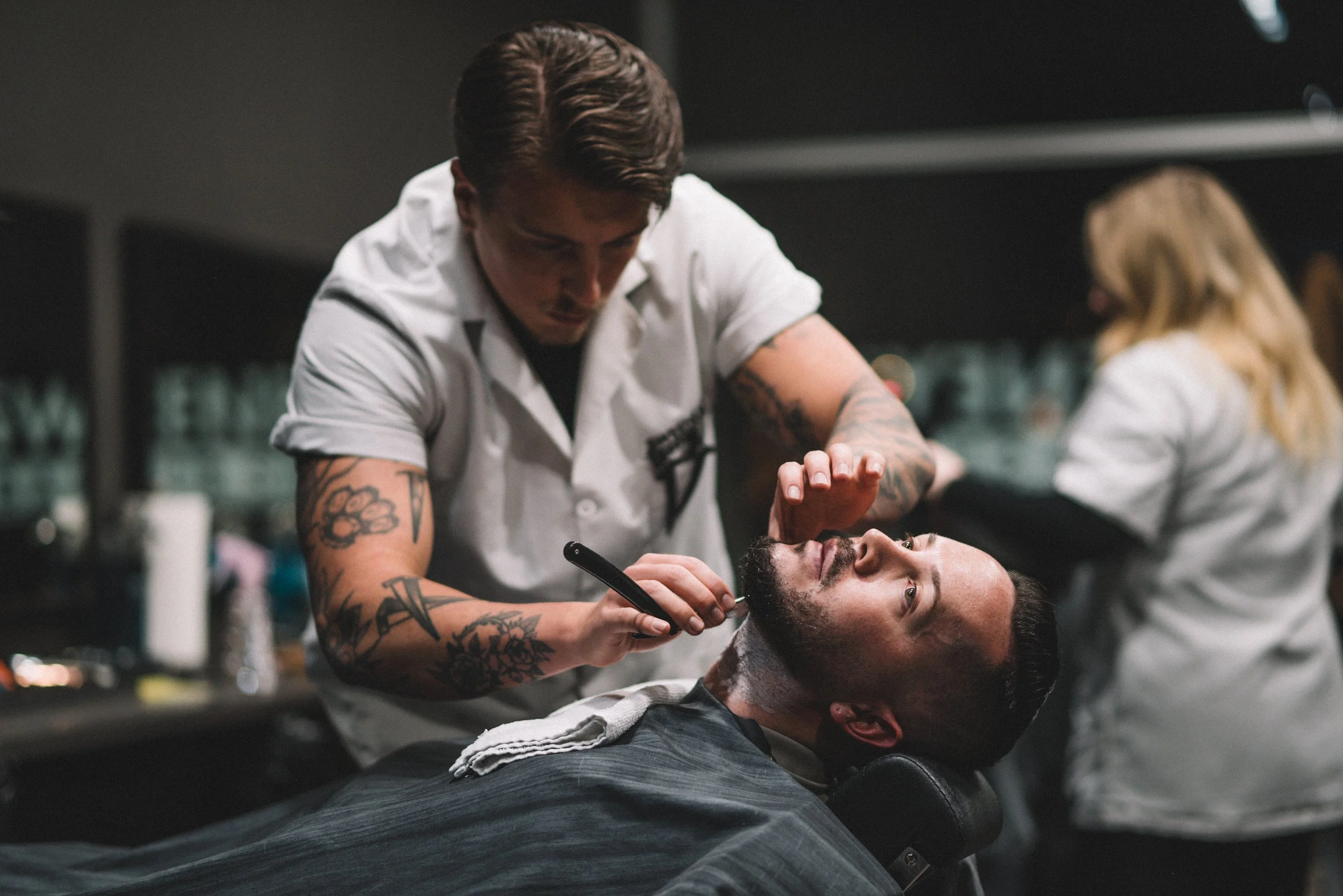 Barber shaving a man's face in a barbershop, with another barber working in the background.