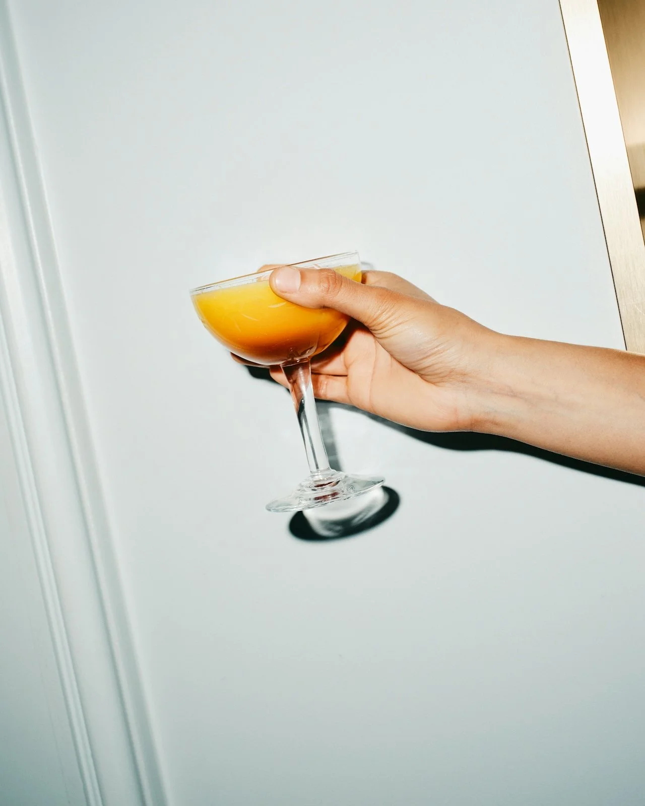 Person holding a cocktail glass filled with orange-colored drink against a plain background.