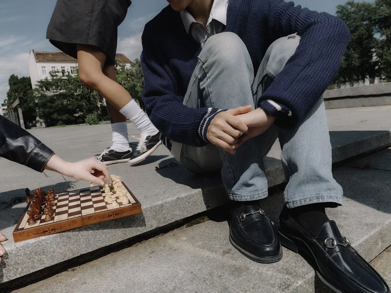 A person sitting on a stone ledge playing chess outdoors while another child plays nearby. The scene is in an open area with buildings and trees in the background.