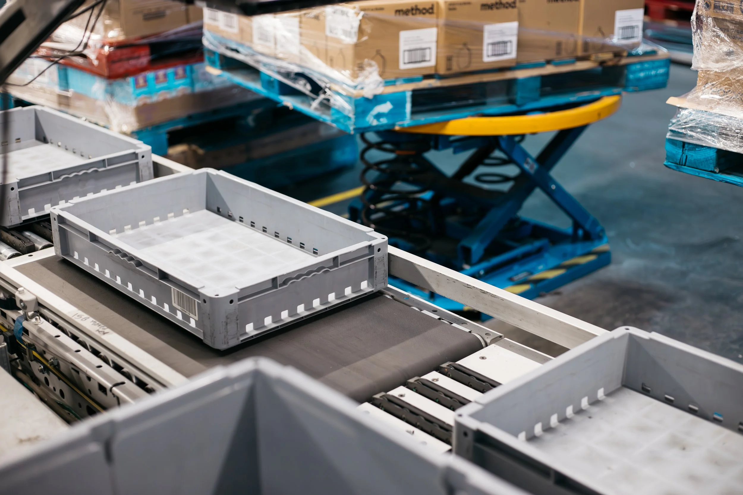 Industrial conveyor belt with white plastic crates in a warehouse or factory setting, with pallets and boxed goods in the background.