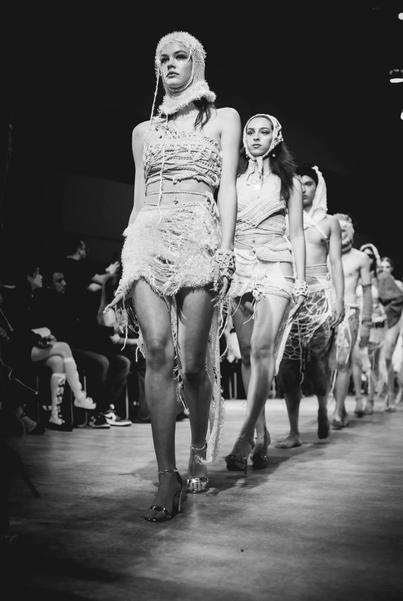 Models walking on a fashion runway wearing distressed and textured clothing in a fashion show, with audience members seated on the side.
