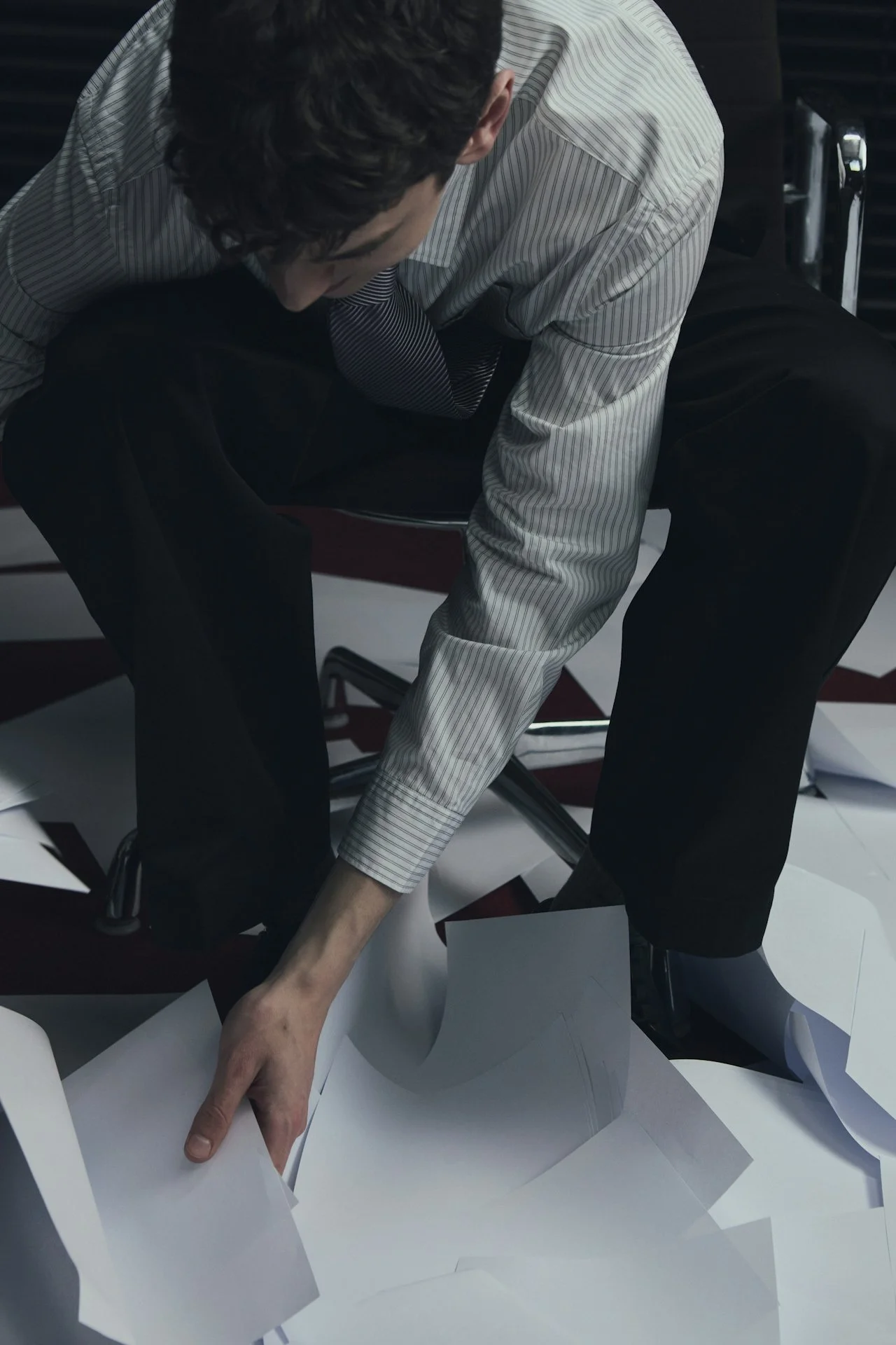 A man in dress shirt and black pants sitting on a chair, surrounded by scattered papers on the floor, reaching to pick up a sheet of paper.