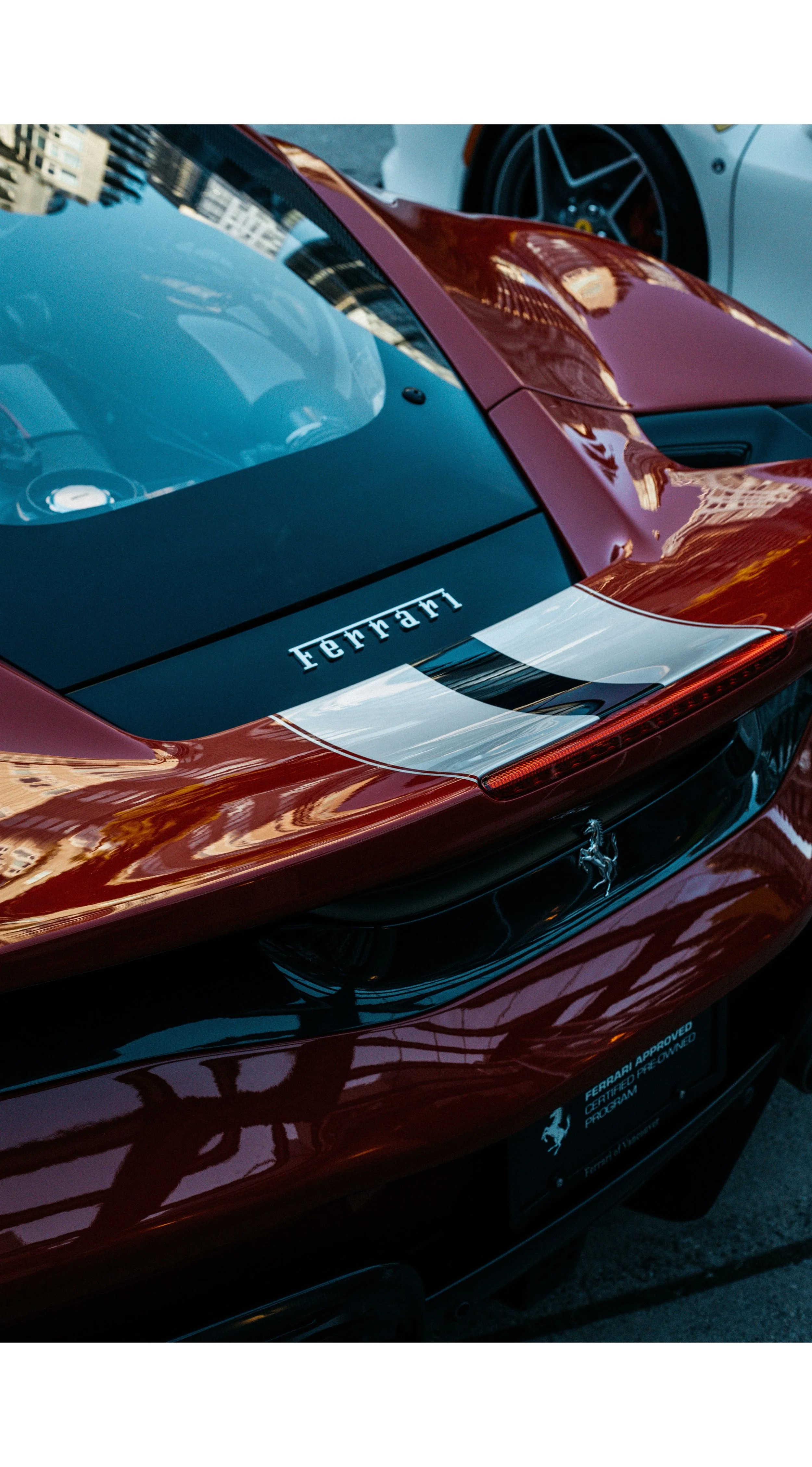 Close-up of the rear of a red Ferrari sports car with a black rear window, a silver stripe on the trunk, and the Ferrari logo. Reflections of buildings and the sky are visible on the car's surface.