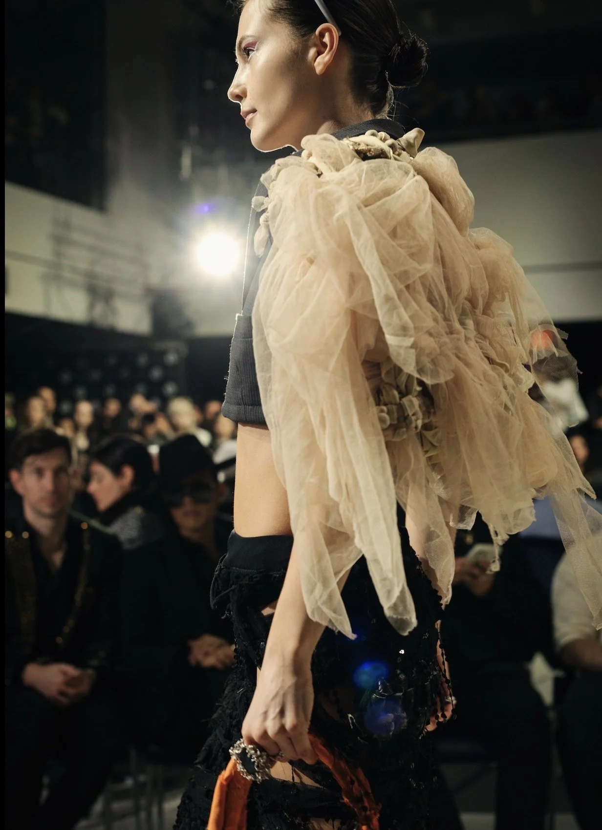A female fashion model walking on runway with beige tulle ruffle detail top, black shorts, and accessories, with audience seated in the background.