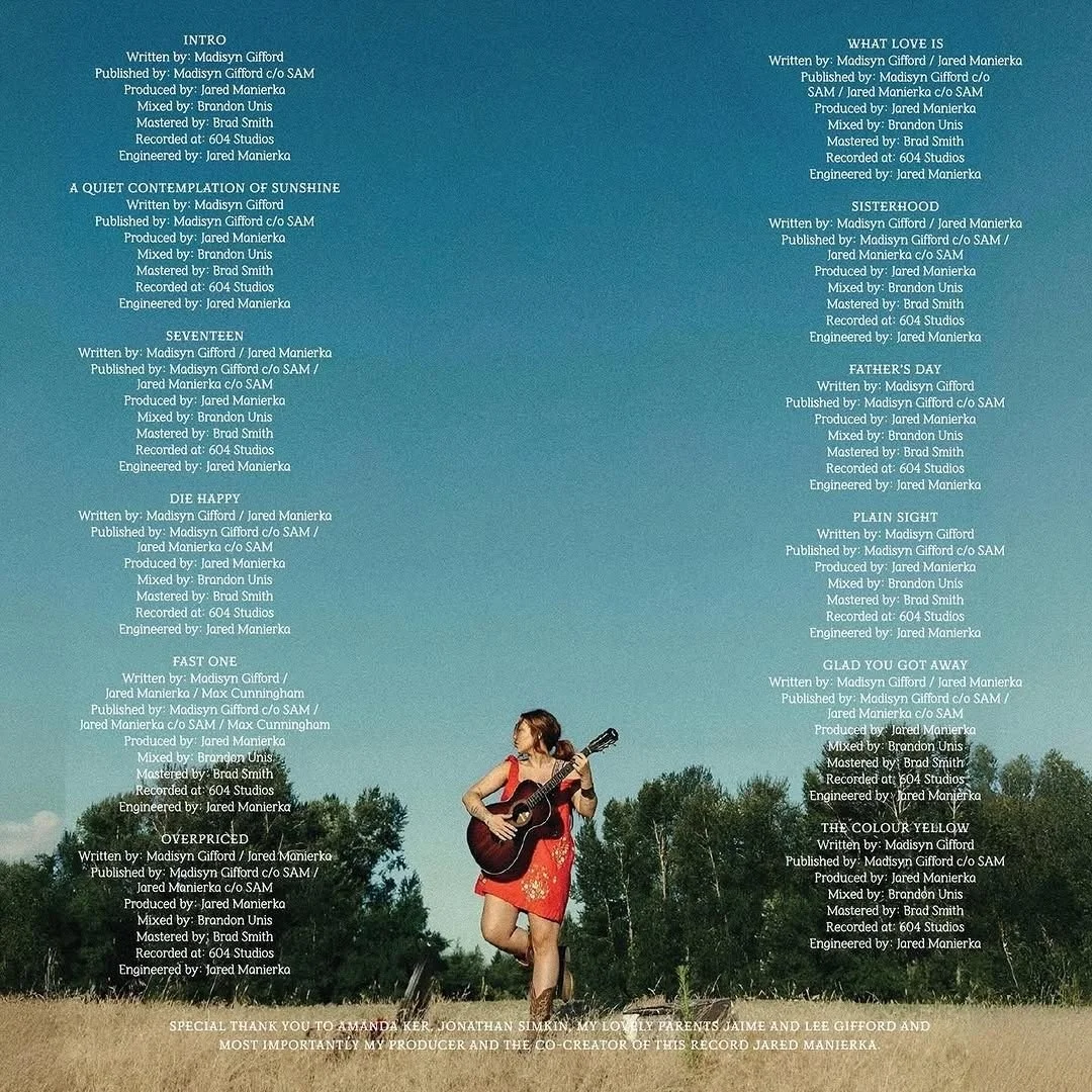 A woman in a red dress playing an acoustic guitar outdoors in a grassy field with trees in the background under a clear blue sky.