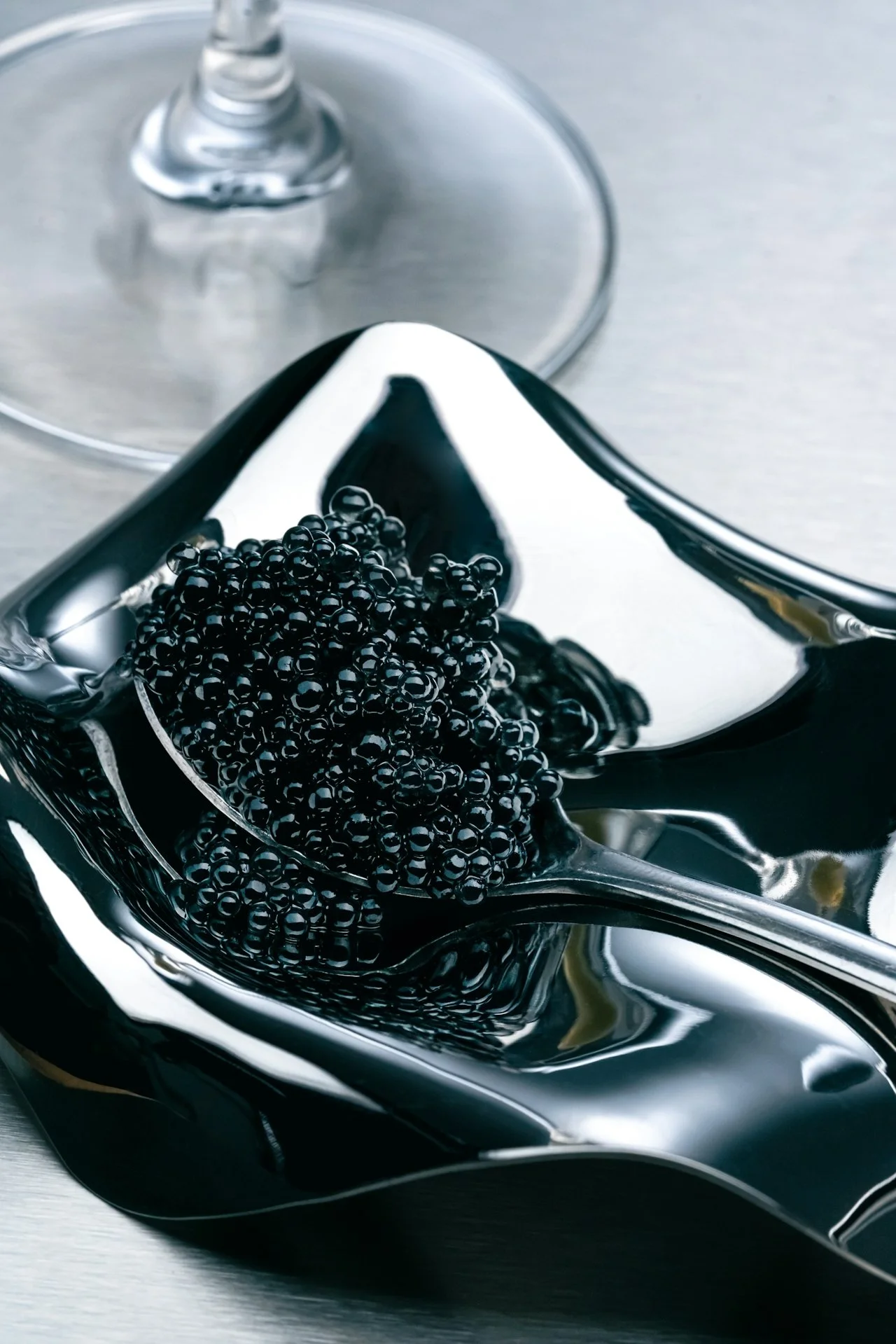 A silver spoon filled with black tapioca pearls resting on a metallic surface, with a glass of wine in the background.