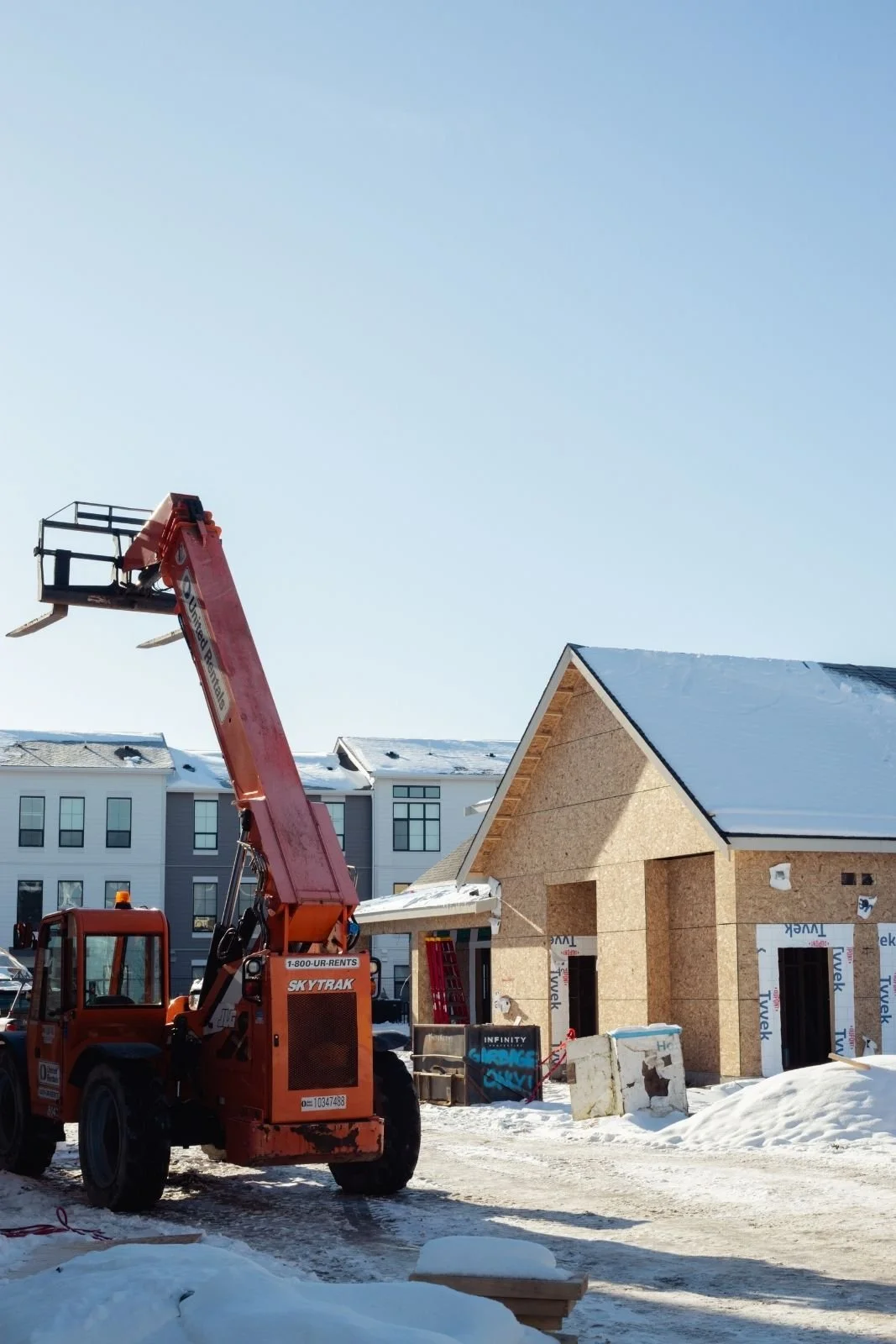 Construction site with a red crane in front of a partly completed house with snow on the ground and a clear blue sky.