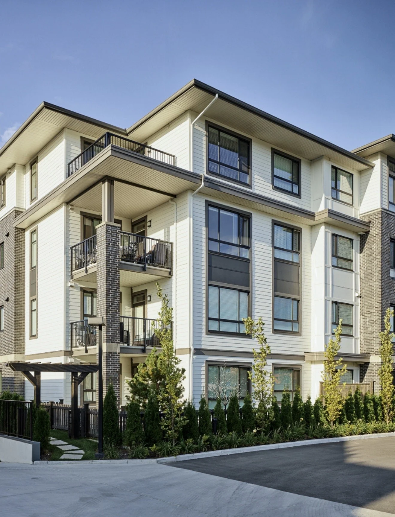 Exterior view of a modern multi-story apartment building with white siding, black window frames, and balconies, surrounded by landscaped greenery under a clear blue sky.