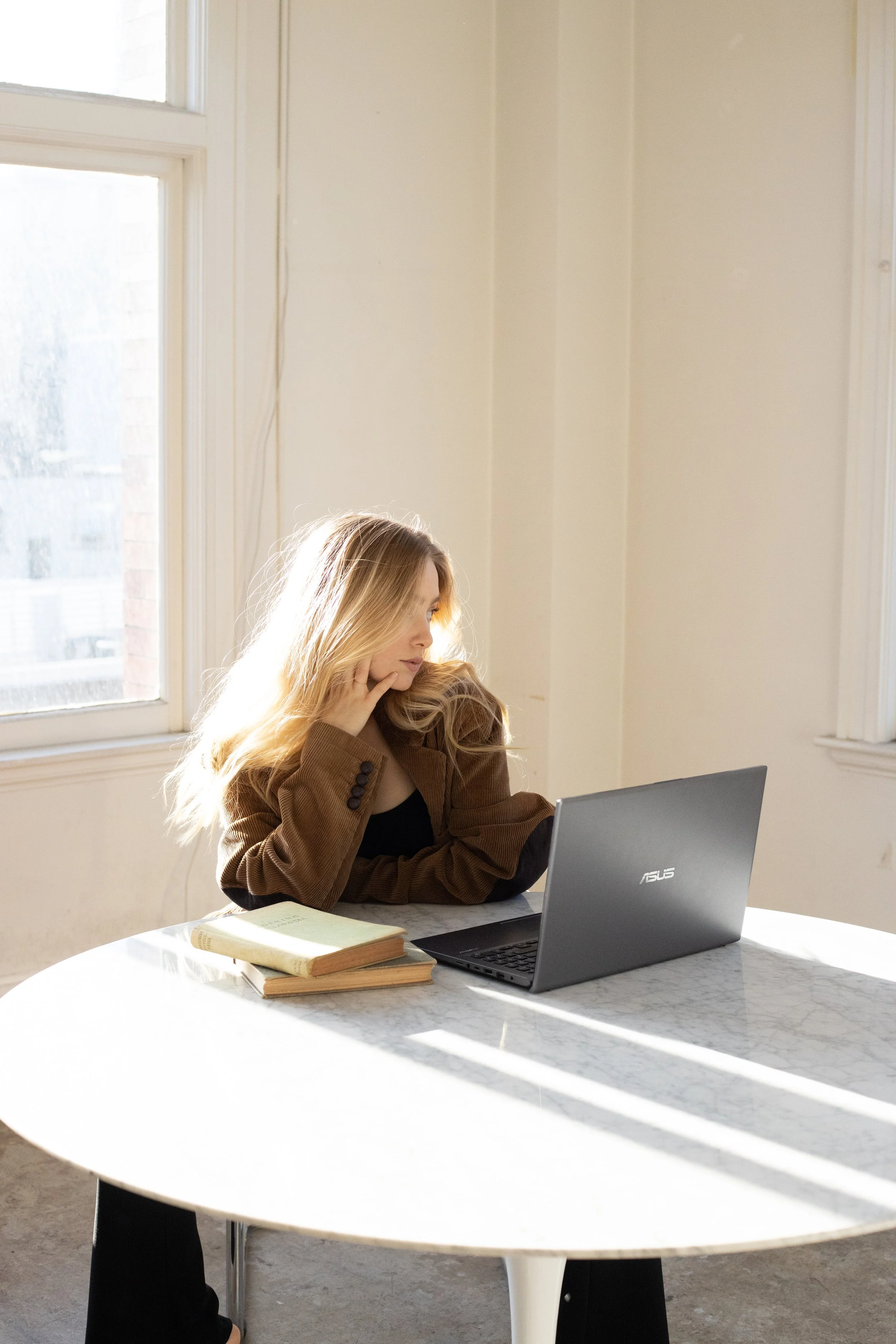 A young woman with blonde hair sits at a round marble table, looking at her laptop with a stack of books beside her. She is inside a bright room with large windows.