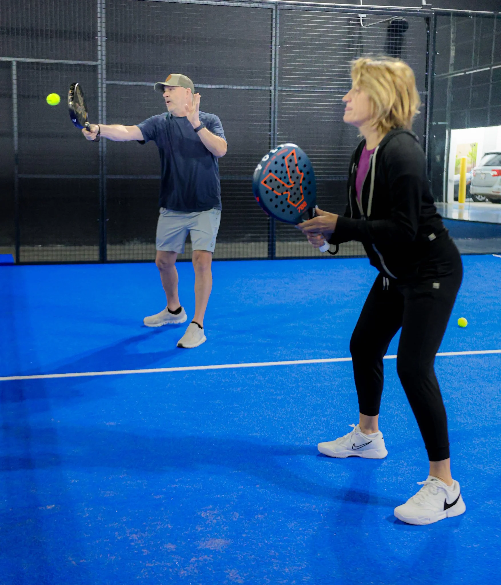 Two people playing indoor padel on a blue court, with one person hitting the ball and the other holding a Volt Padel racquet. 