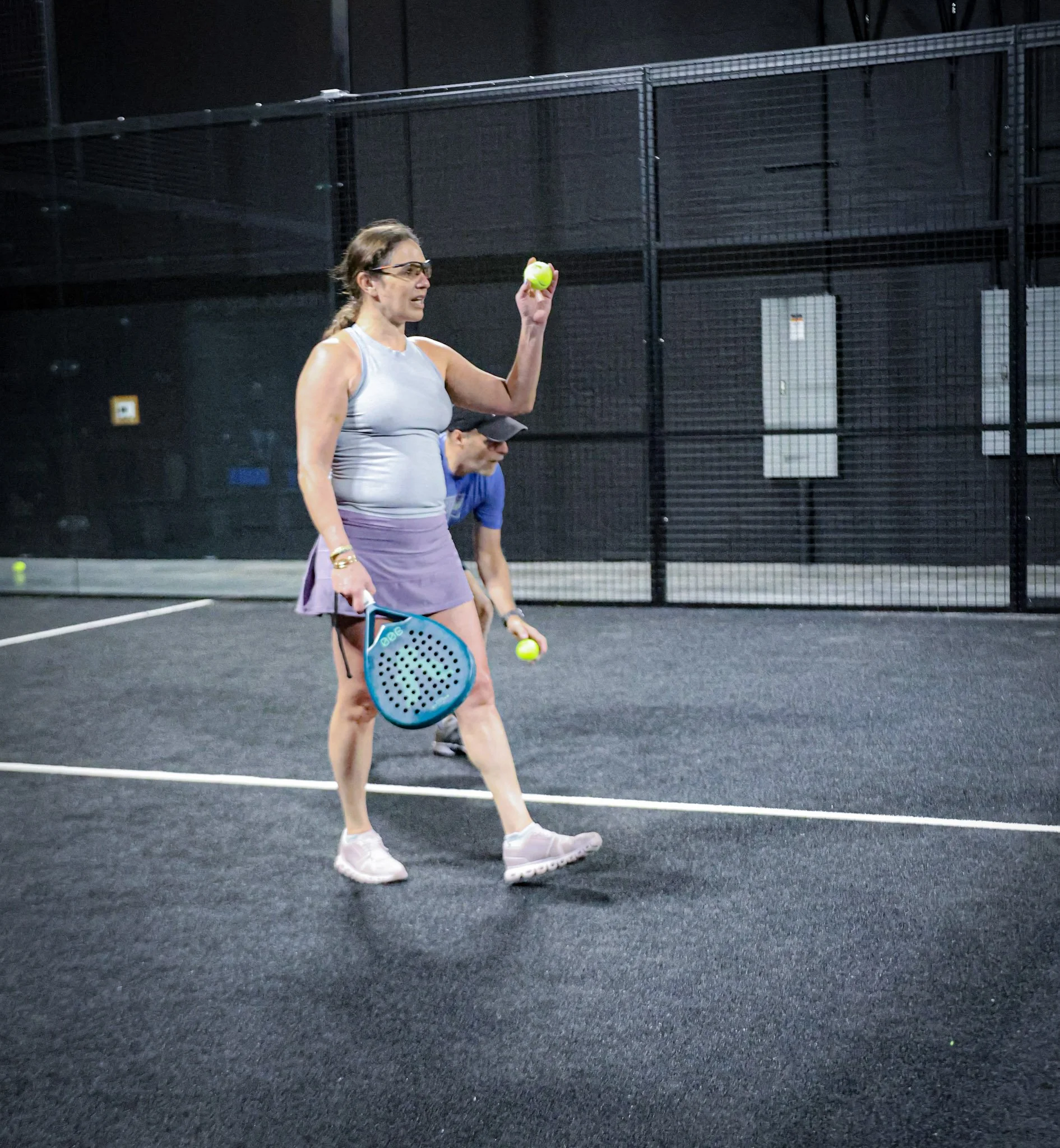 A woman wearing glasses, a tank top, and shorts, standing on a padel court, holding a padel ball in her right hand and a padel racquet in her left hand. She is looking at the ball with a focused expression. 