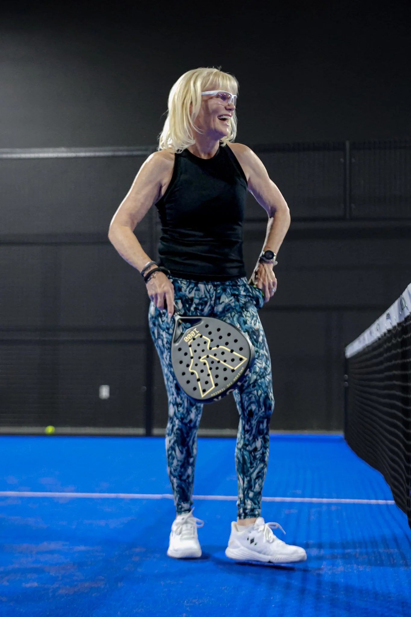 A woman with blonde hair wearing sunglasses, a black athletic top, patterned leggings, and white sneakers holding padel racquets on an indoor blue padel court at a profitable padel club.