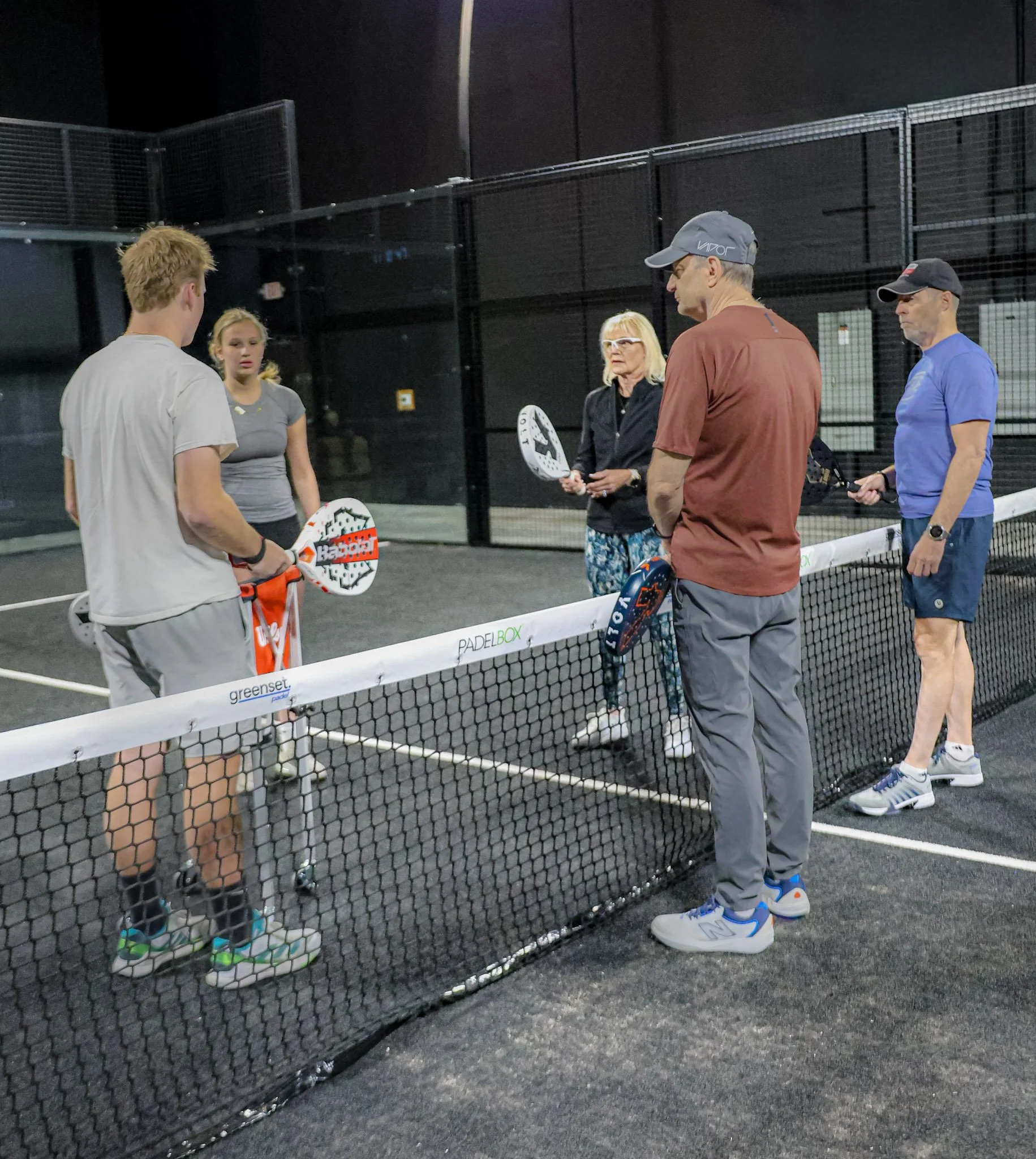 Group of five people standing on a padel tennis court, three men and two women talking, with two padel racquets visible, one held by a woman and the other held by a man, and a child in padel shoes standing near the net.