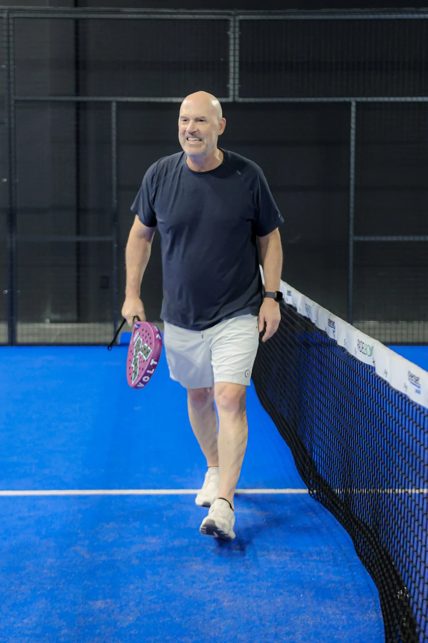 A man with a shaved head wearing a dark t-shirt and white shorts walking on a blue padel court holding a teardrop-shaped padel racquet.