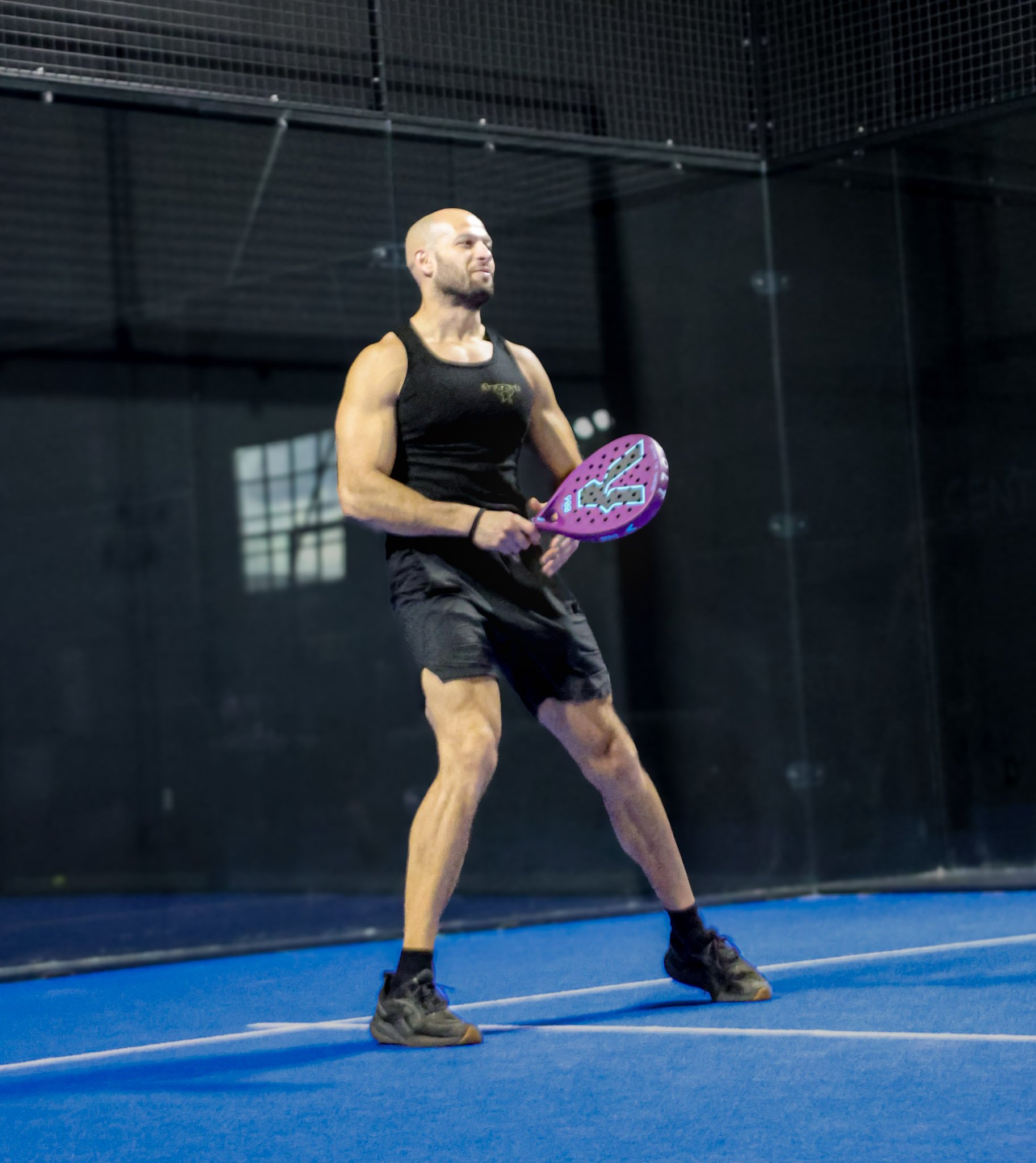 A man holding a padel racquet stands on a blue padel court with black walls and a metal grid ceiling, dressed in black athletic wear.