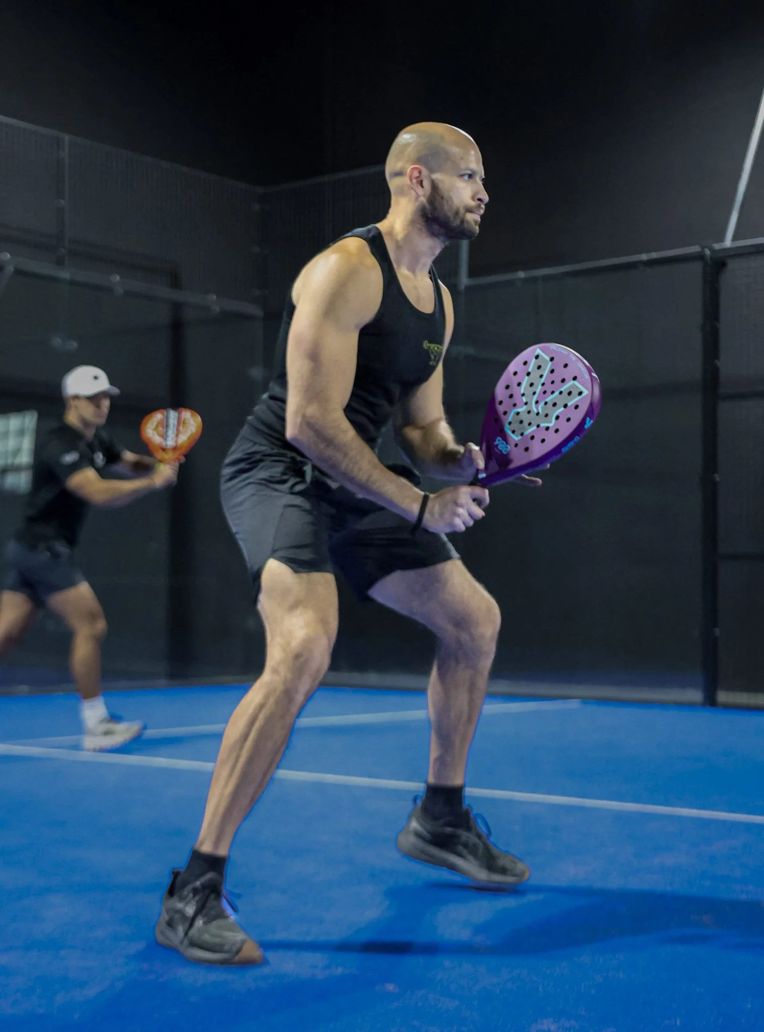 A man playing padel on an indoor padel court, holding a padel racquet, wearing athletic attire including a black tank top, shorts, and sneakers. In the background, another player holds a teardrop-shaped padel racquet.