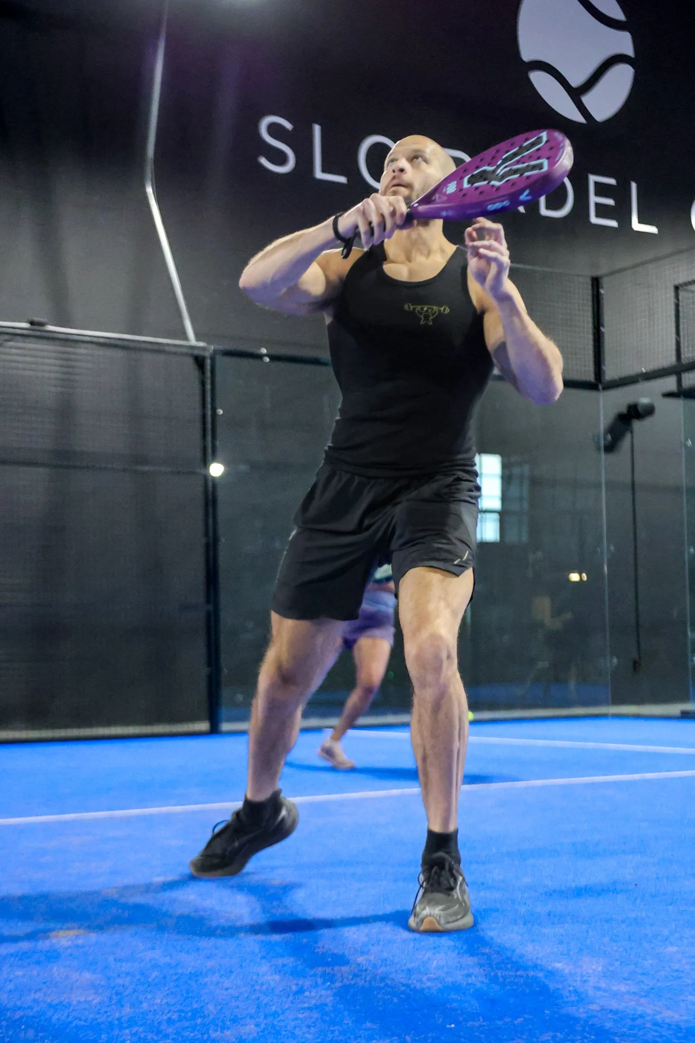Man in black tank top and shorts holding a purple padel racquet in a profitable padel club with a blue floor and black wall, reflected in a mirror.