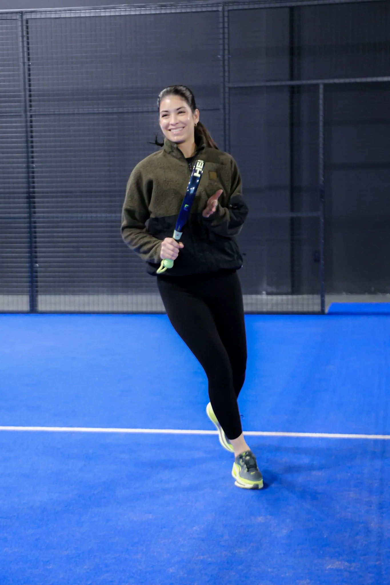 Woman running on blue indoor padel court, holding padel racquet, smiling.