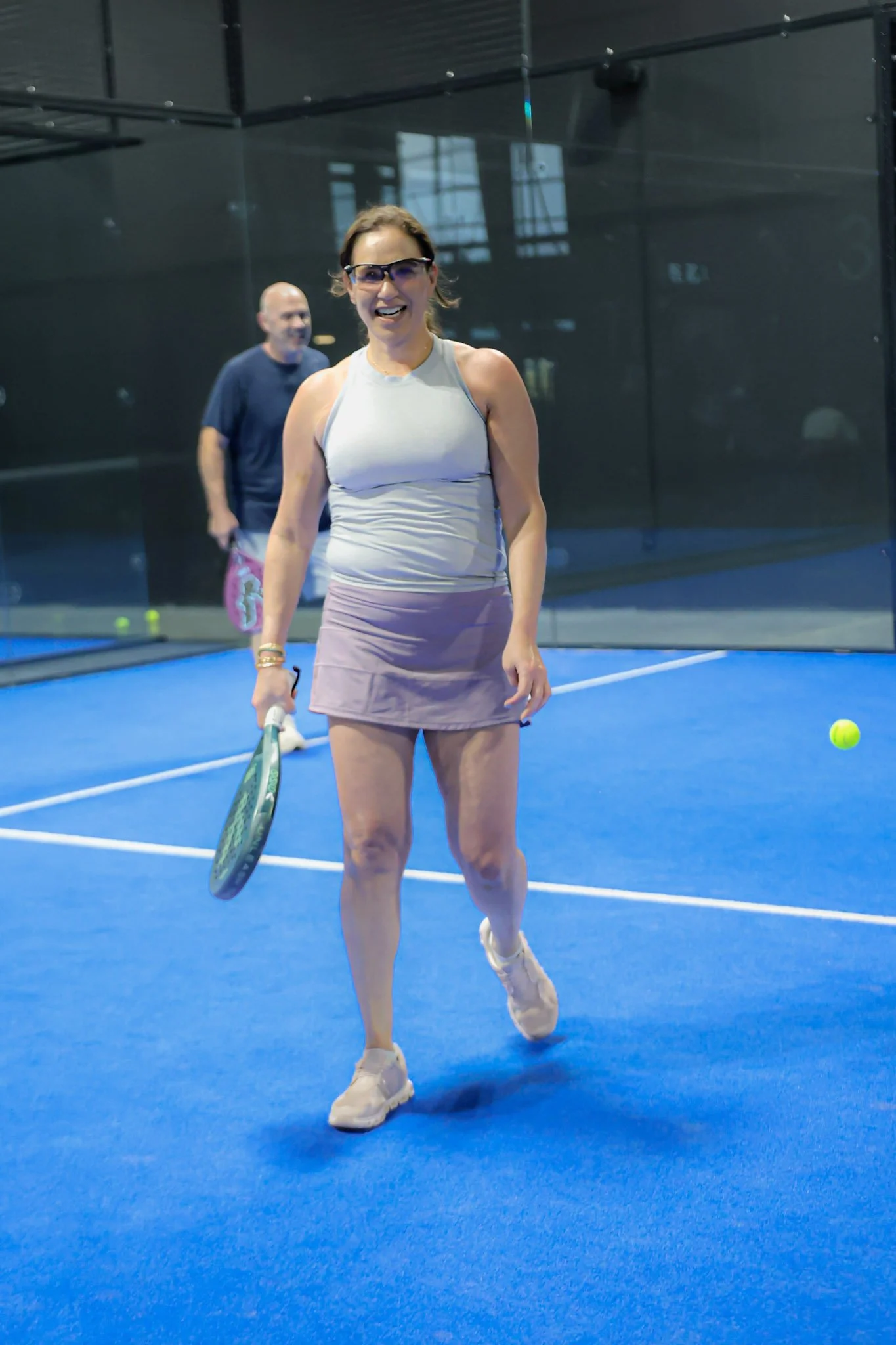 A woman on a padel court holding a padel racket, smiling, with a man in the background, all inside an indoor padel club.
