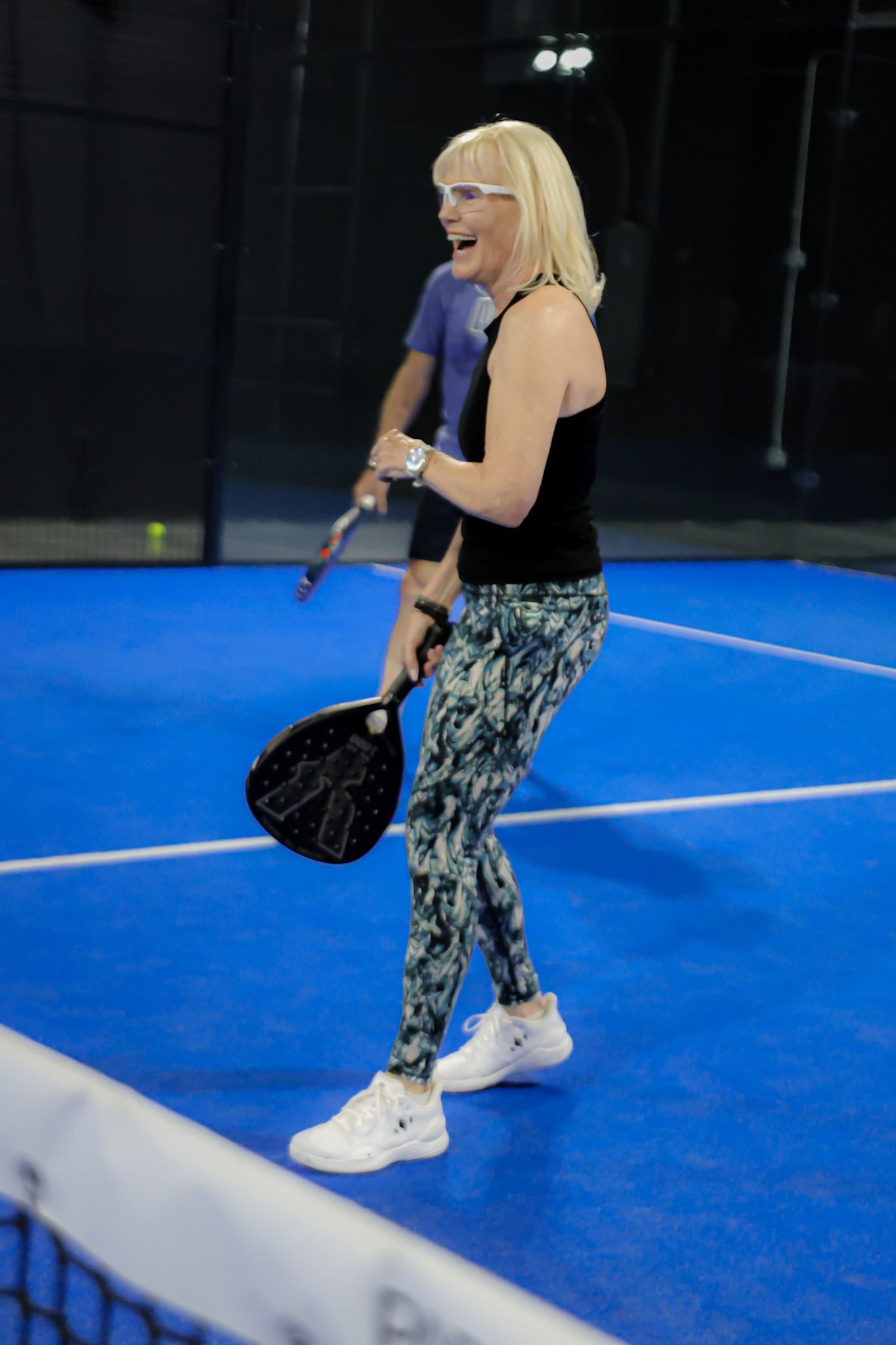 A woman playing padel, holding a padel racquet and a ball, wearing glasses, a black tank top, patterned leggings, and white sneakers, on a blue padel court with a net in the foreground.