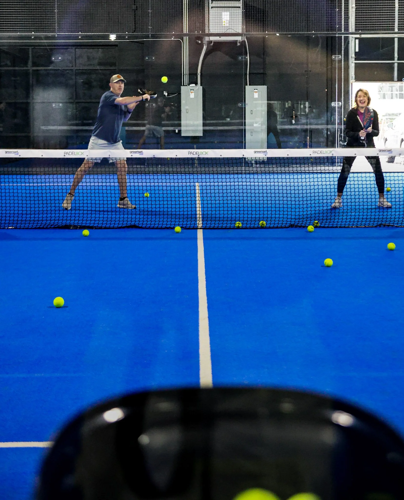 An indoor padel court with a man and woman playing padel, seen through the glass wall. The court has a blue playing surface and yellow padel balls scattered across it, with netting and black walls in the background.