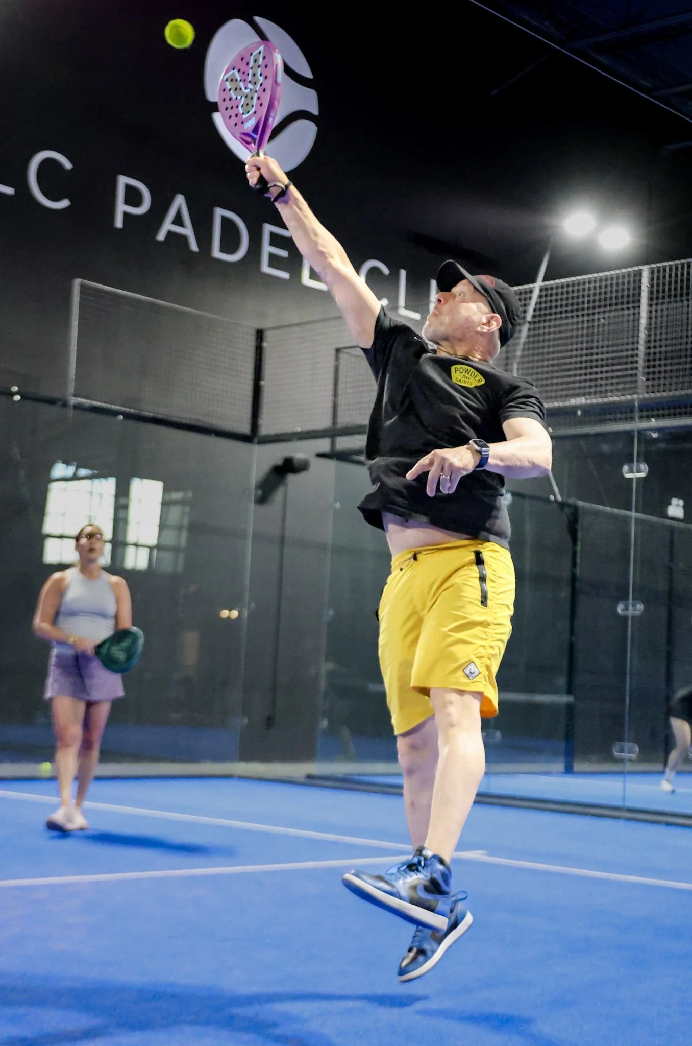 A man is playing padel on an indoor padel court, hitting a ball with a padel racquet, with a woman standing in the background holding a padel racquet and ball.