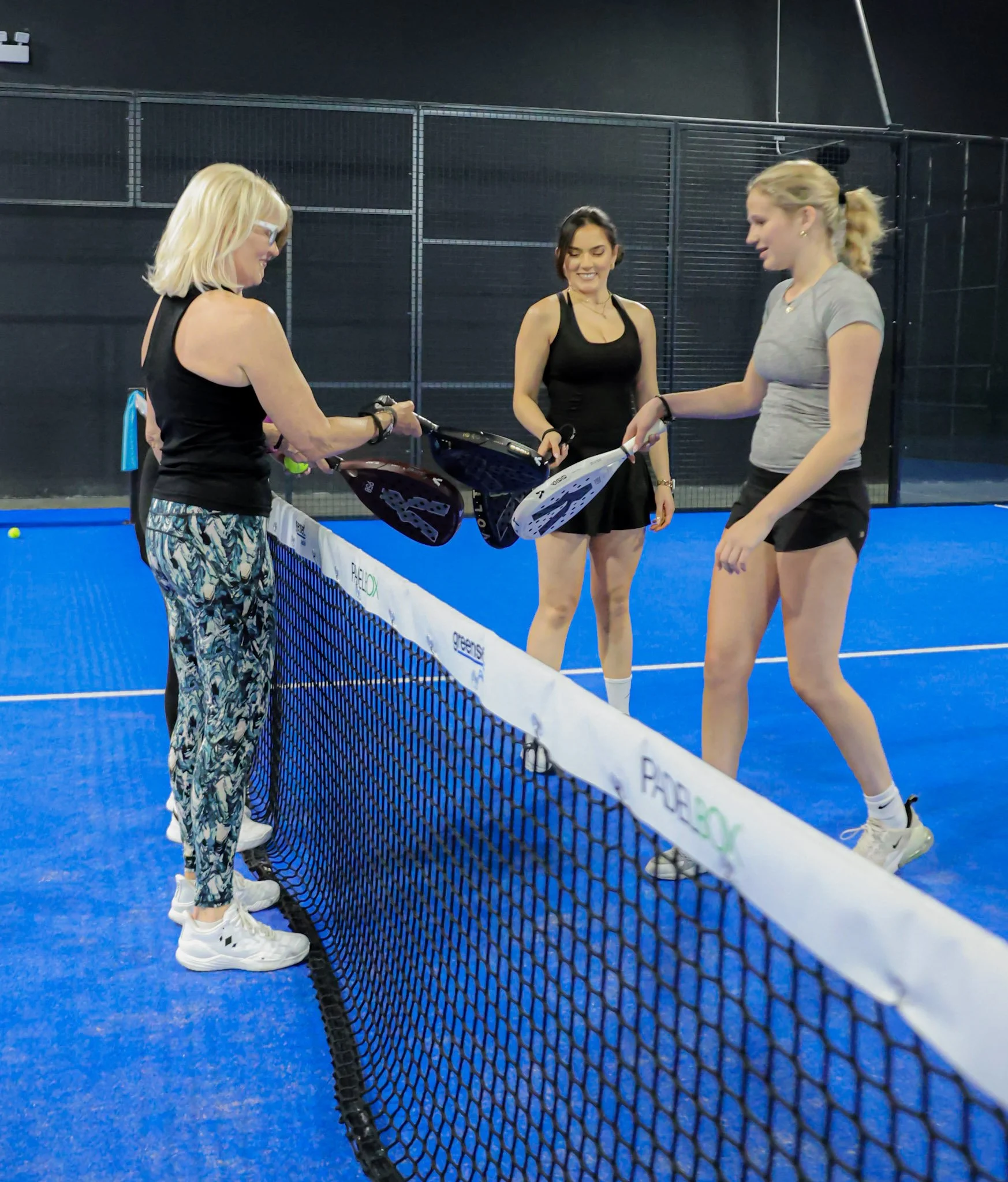 Three women inside an indoor padel court, two of whom are exchanging a handshake, with a net separating them. All women are dressed in tennis dress or tennis skirts, and the padel court has a blue surface.