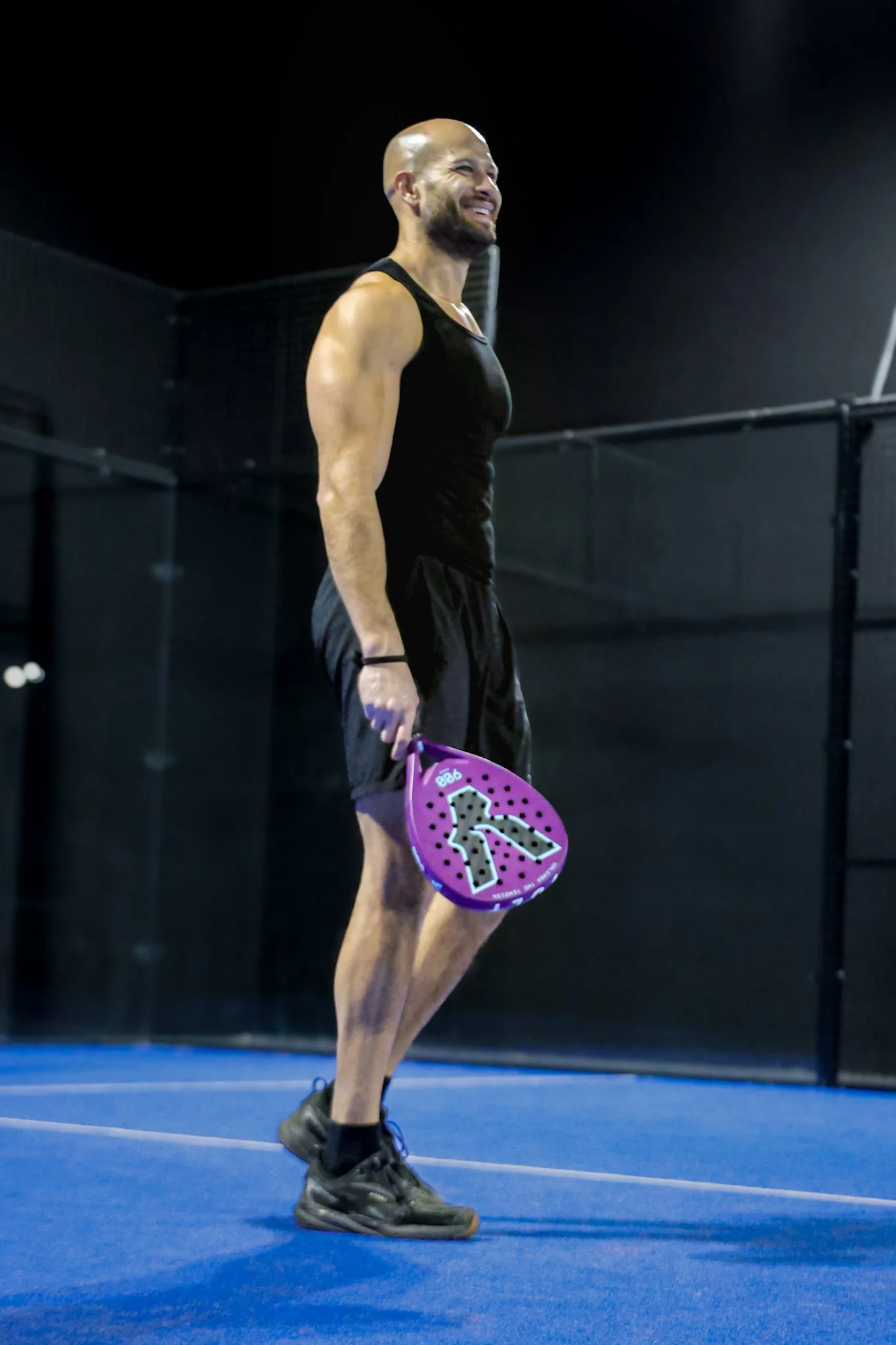 A man in black athletic attire and sneakers holding a Volt Padel while standing on a blue padel court, smiling.