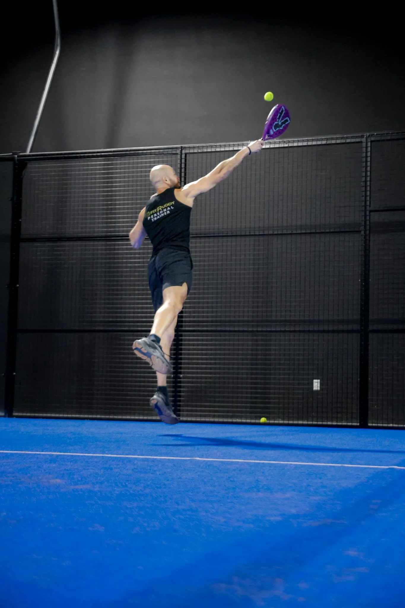 A man playing padel indoors, hitting a green tennis ball with a purple paddle over a black safety net, on a blue padel court.