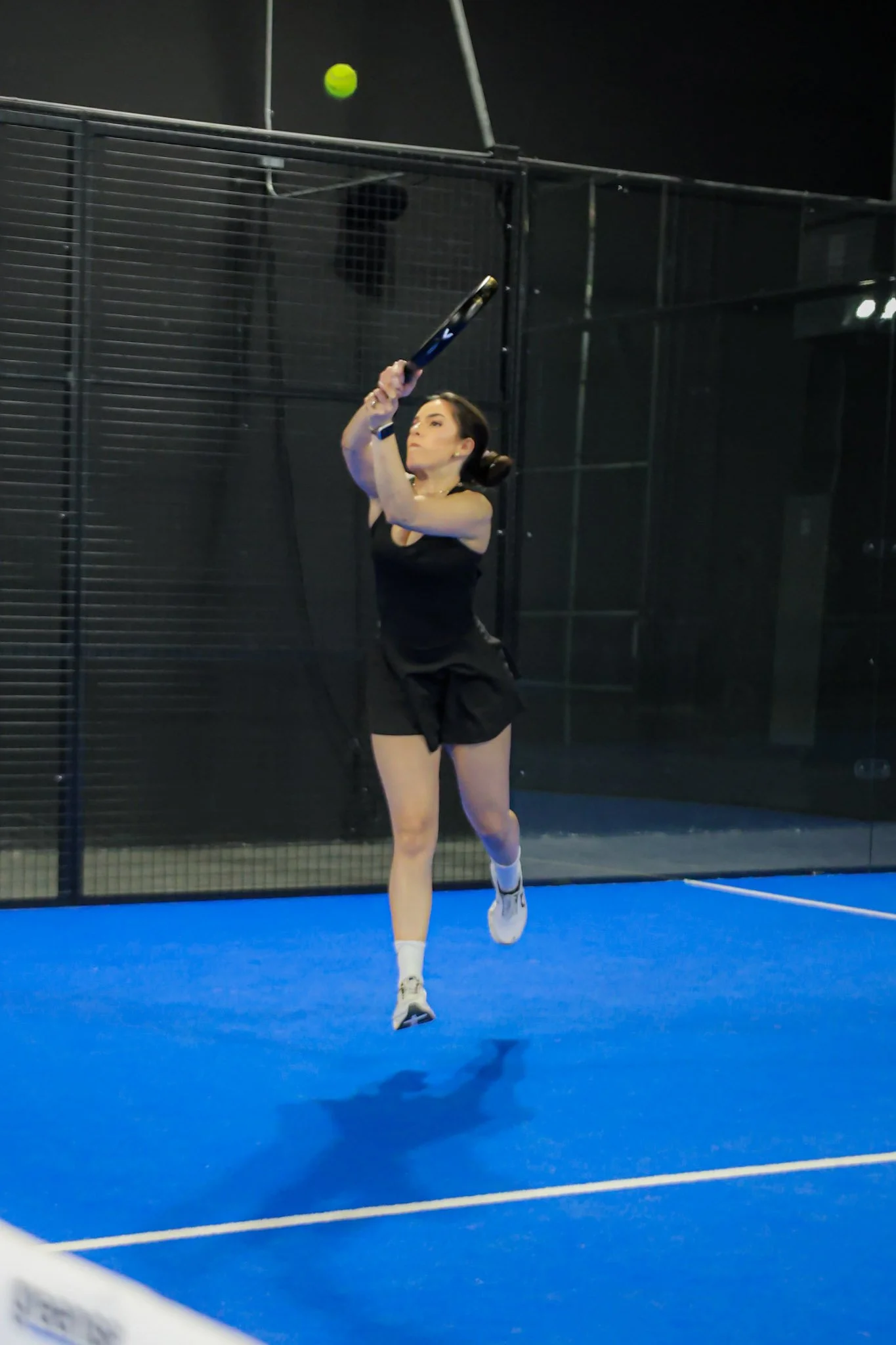 A woman playing padel on an indoor padel court, reaching up to hit a green padel ball with a black padel racquet.