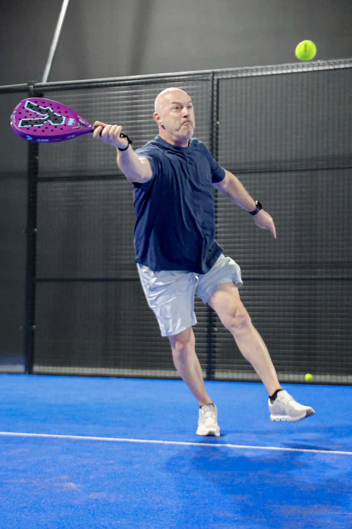 A man playing padel tennis on an indoor padel court, preparing to hit a yellow ball with a purple padel racquet.
