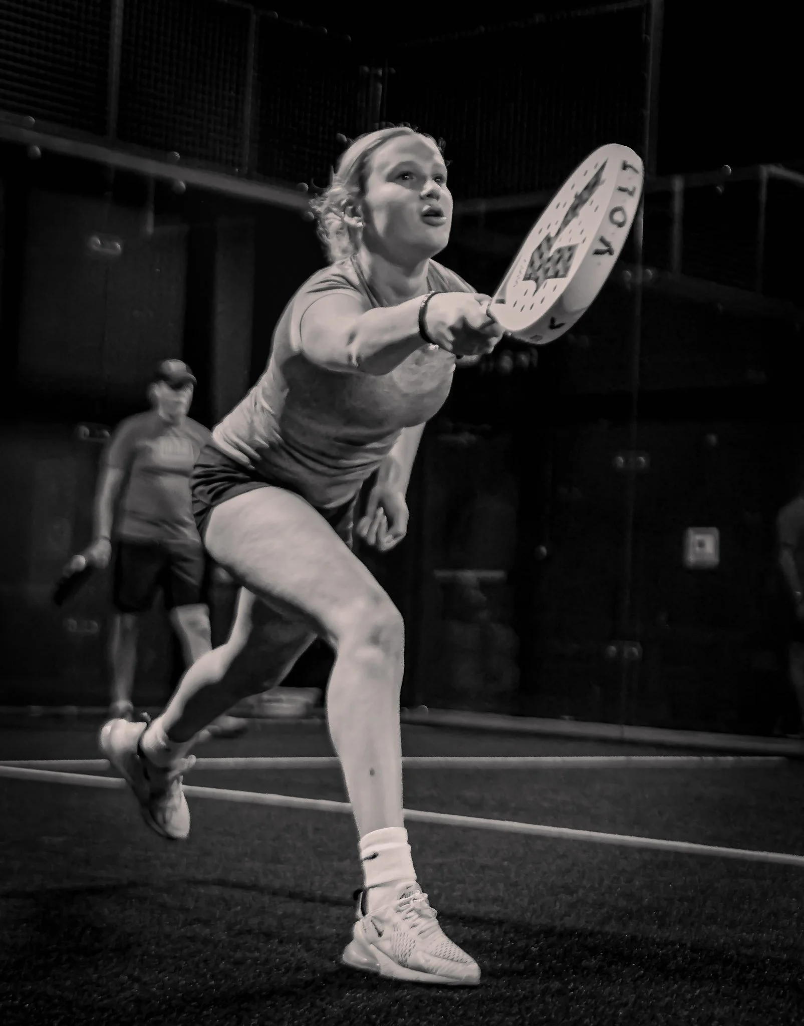 A female padel player in action, reaching out with her padel racquet to hit a ball, on an indoor court with a net in the background.