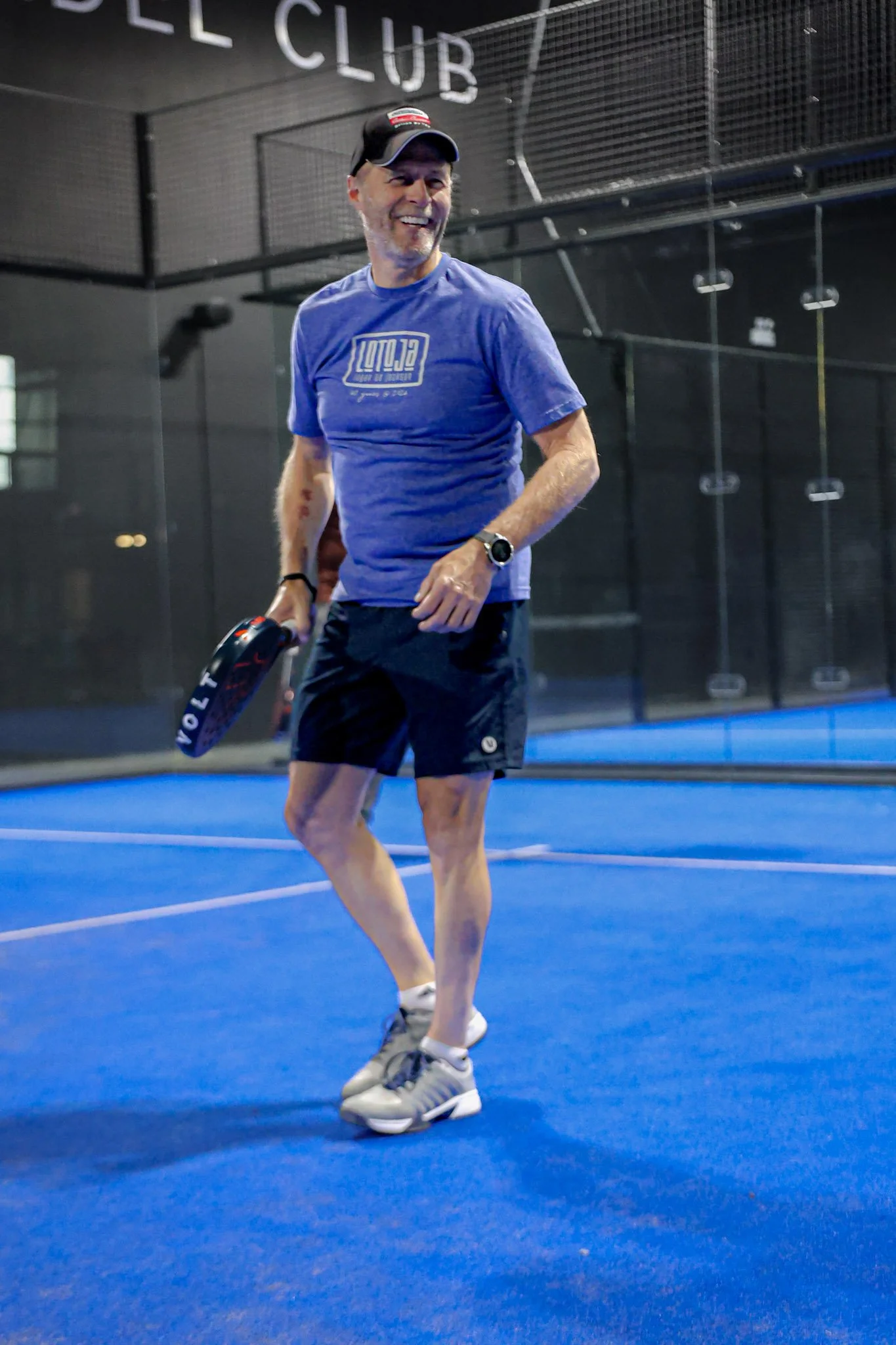 A middle-aged man with a beard, wearing a blue t-shirt, black shorts, gray athletic shoes, a black baseball cap, and a smartwatch, smiling and holding a padel racquet in an indoor padel court.