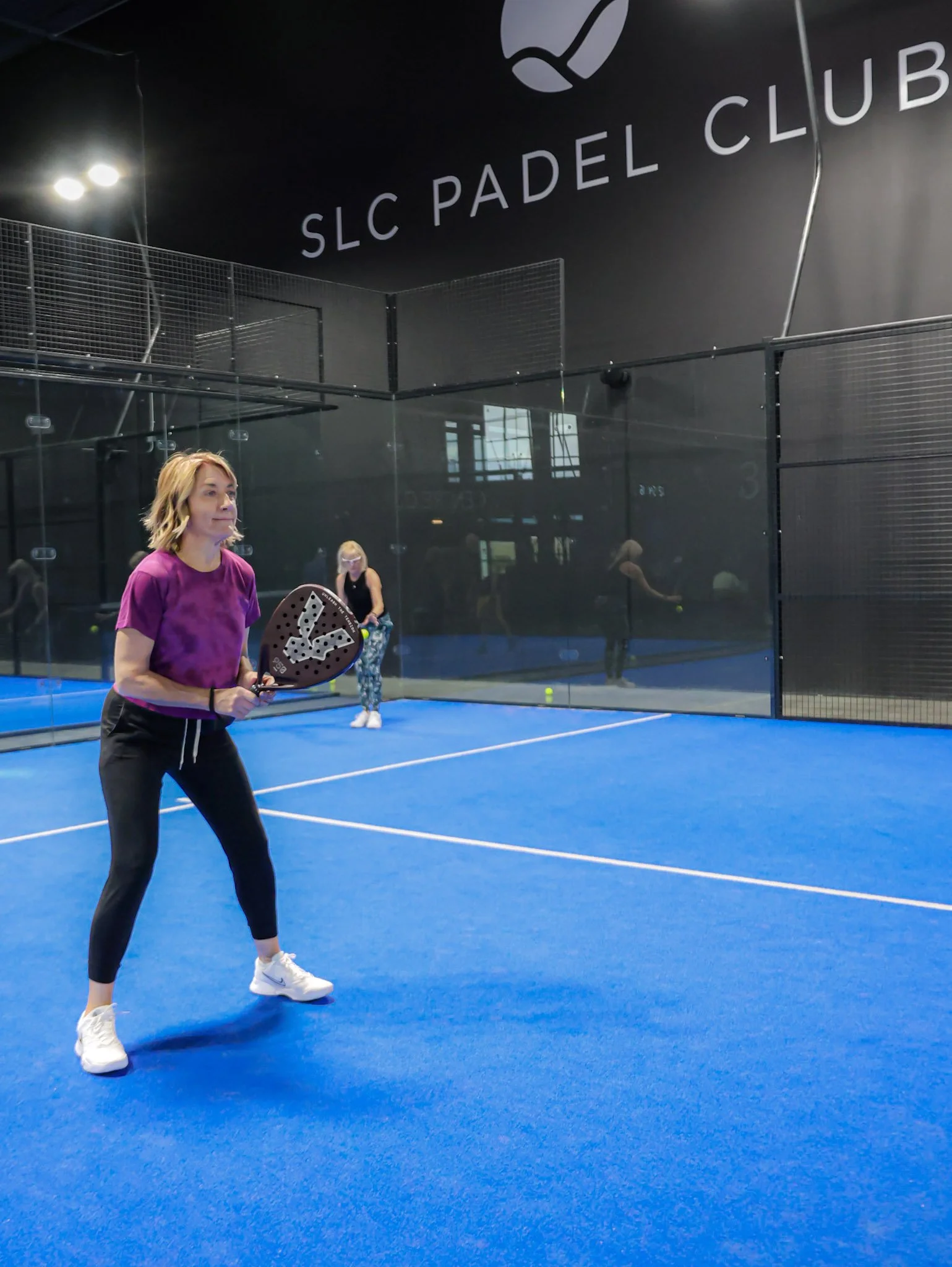 A woman in a purple shirt and black pants playing padel on an indoor padel court with blue flooring, holding a padel racquet, with others playing in the background, and a sign on the wall reading SLC Padel Club.