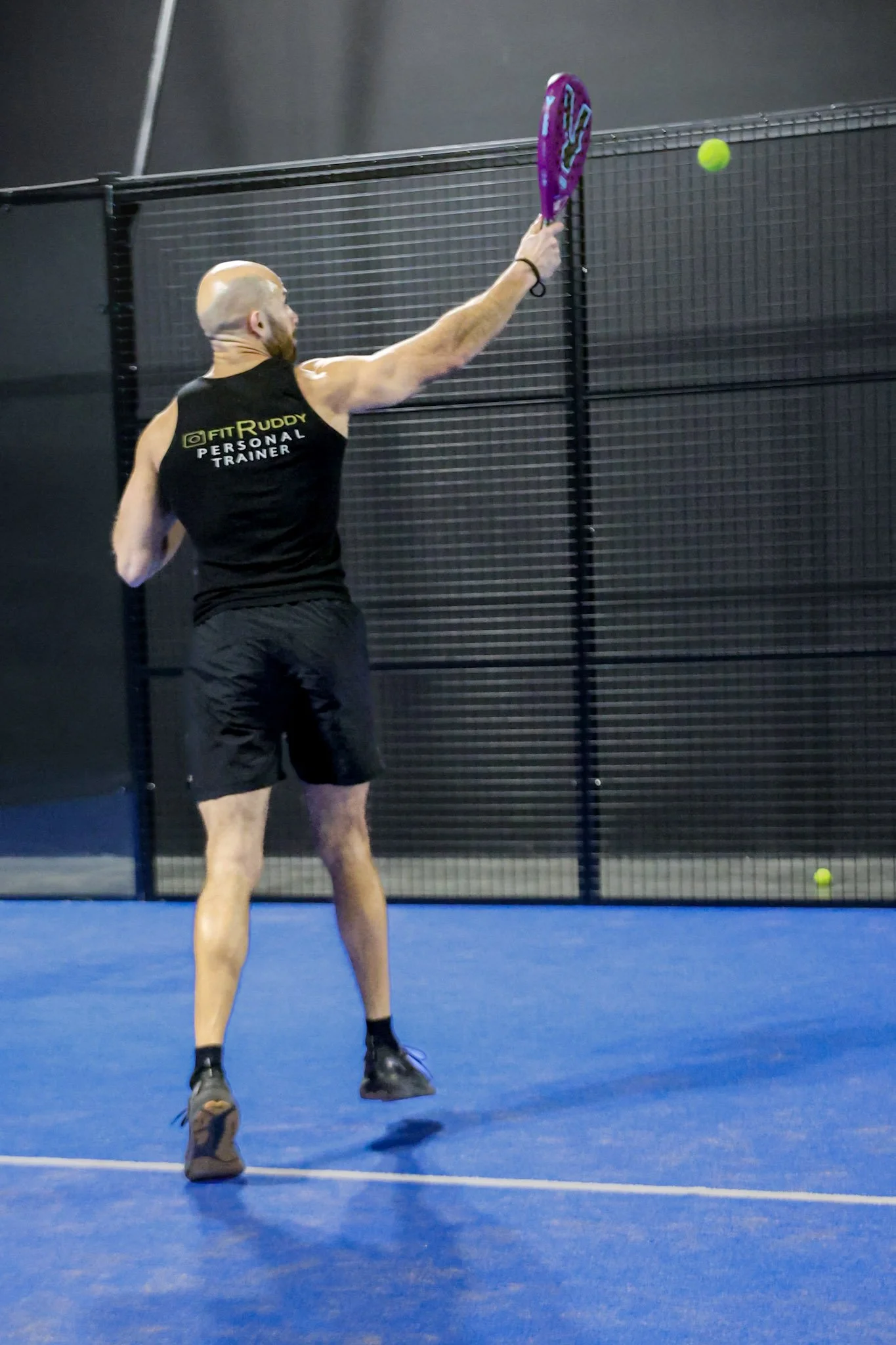 A man is playing padel on a blue padel court with black fencing in the background. He is holding a Volt Padel racquet and about to hit a yellow padel ball.