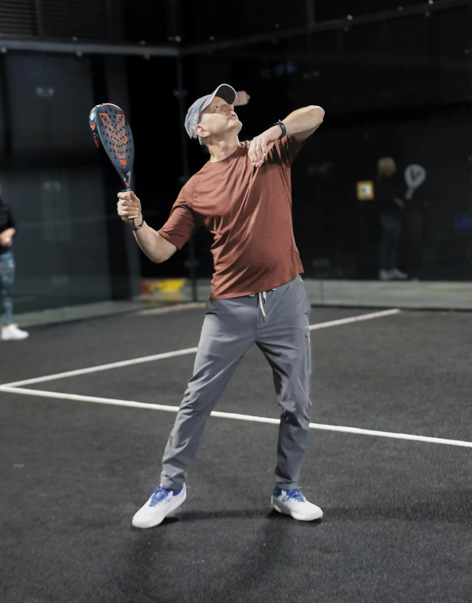 A man in athletic clothing playing padel on an indoor court. He is holding a padel racquet in his right hand, wearing a beige cap, a brown T-shirt, and gray pants with white and blue padel shoes.