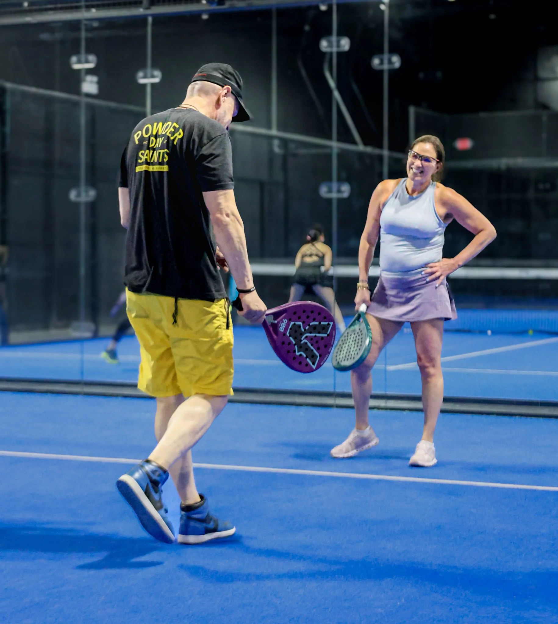 A woman and a man playing pickleball indoors on a blue court. The woman is smiling and wearing a skirt, holding a padel racquet. The man is wearing yellow shorts, with his head down and holding a padel racquet. 